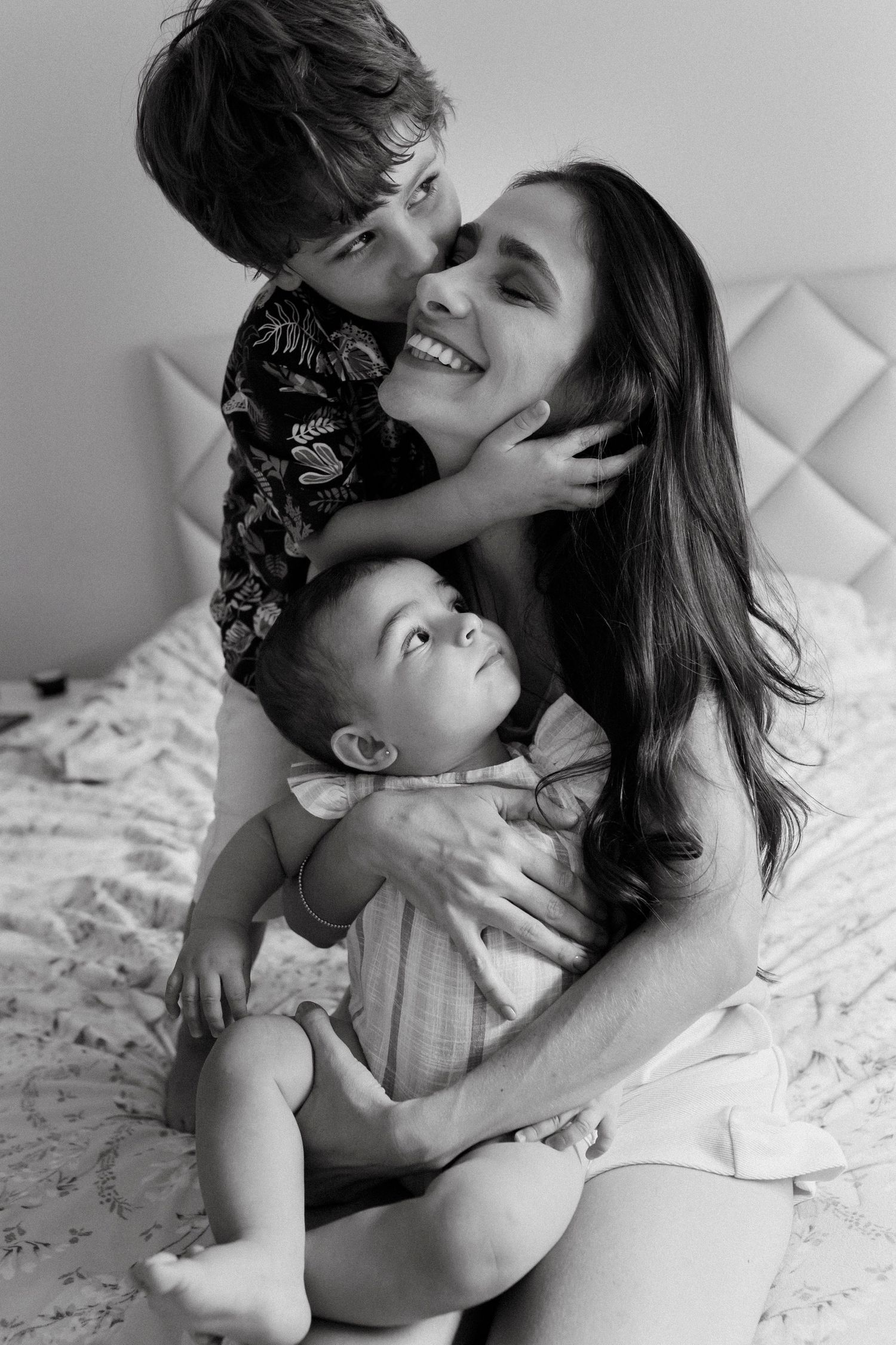A black and white portrait of a mother with two children sharing an intimate moment on a bed.