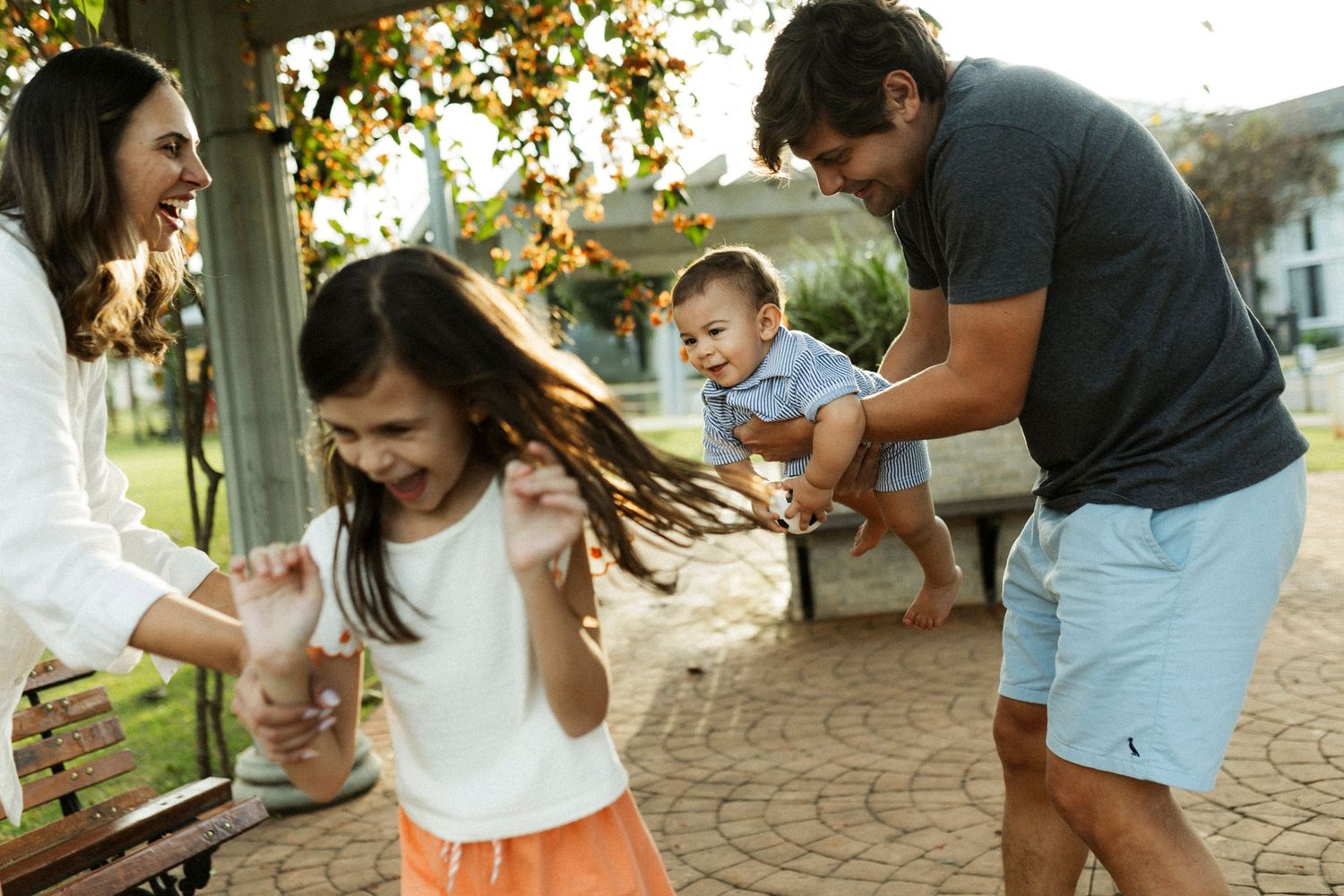 A joyful family plays outdoors, with parents laughing as their young daughter twirls and dad holds a smiling baby.