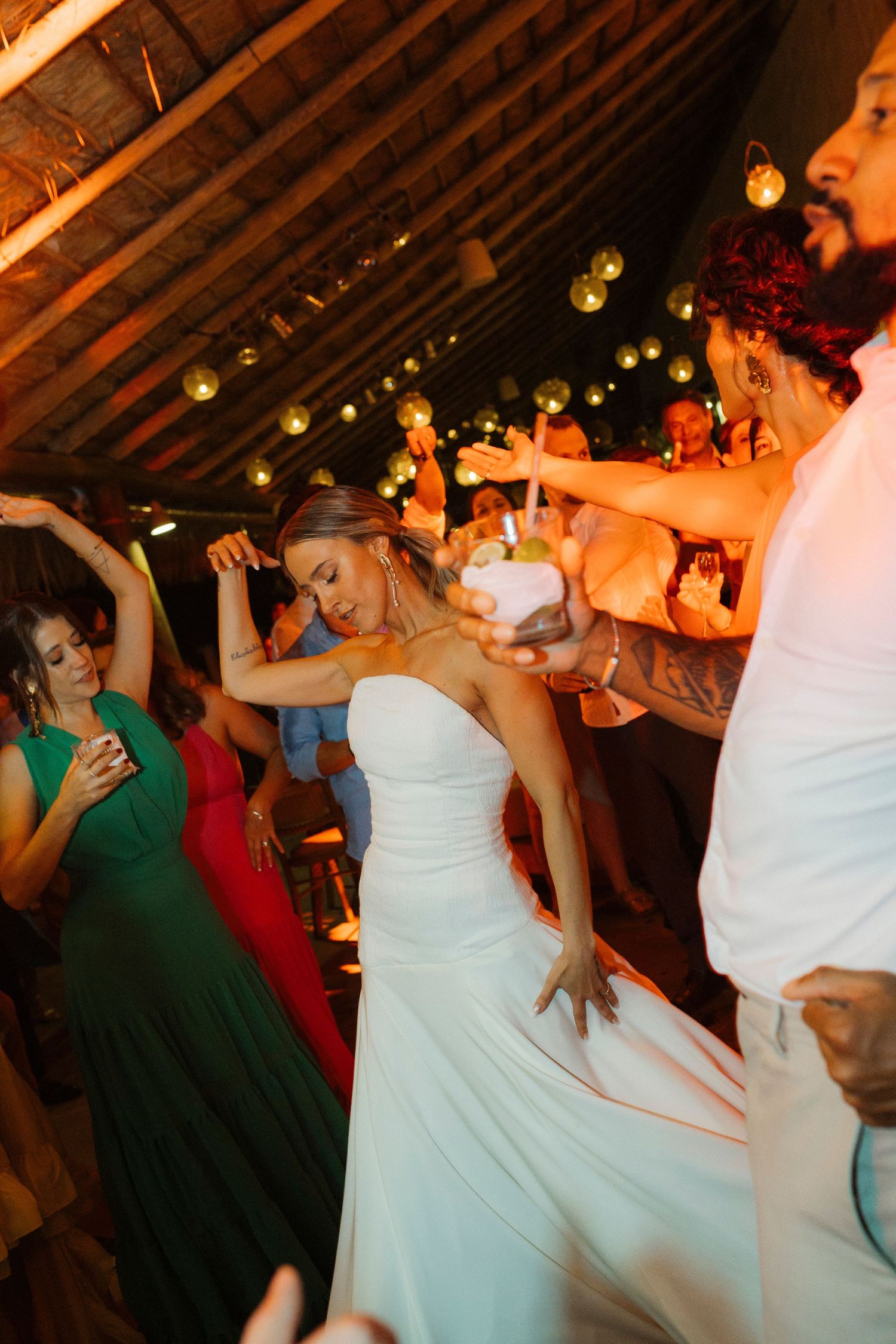 People dancing and celebrating under wooden beams with string lights at a wedding reception.