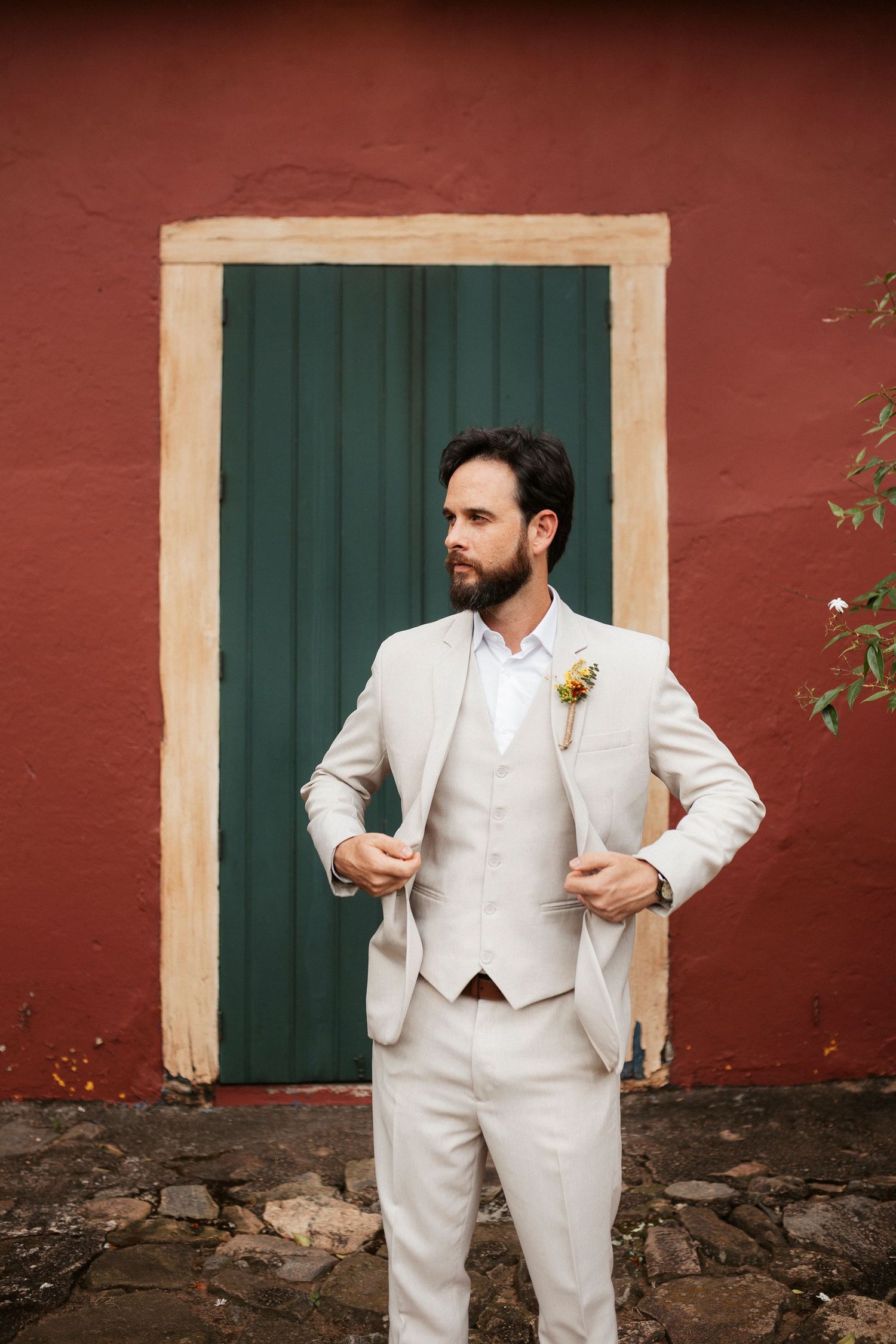 Person in white suit adjusts their attire against a rustic red wall with green shutters.