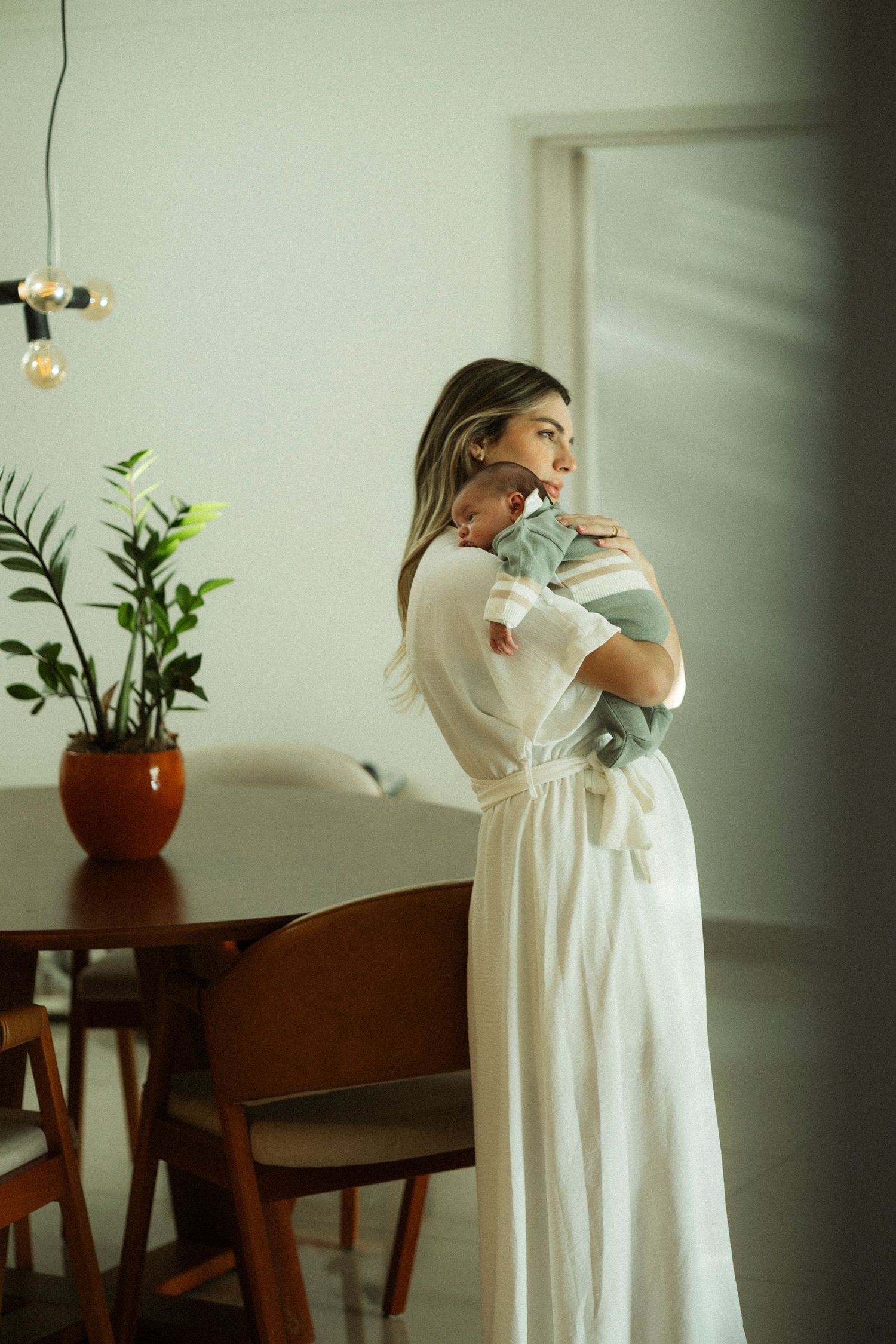 A mother in a white dress tenderly holds her newborn baby in a modern dining room with warm wood furniture.
