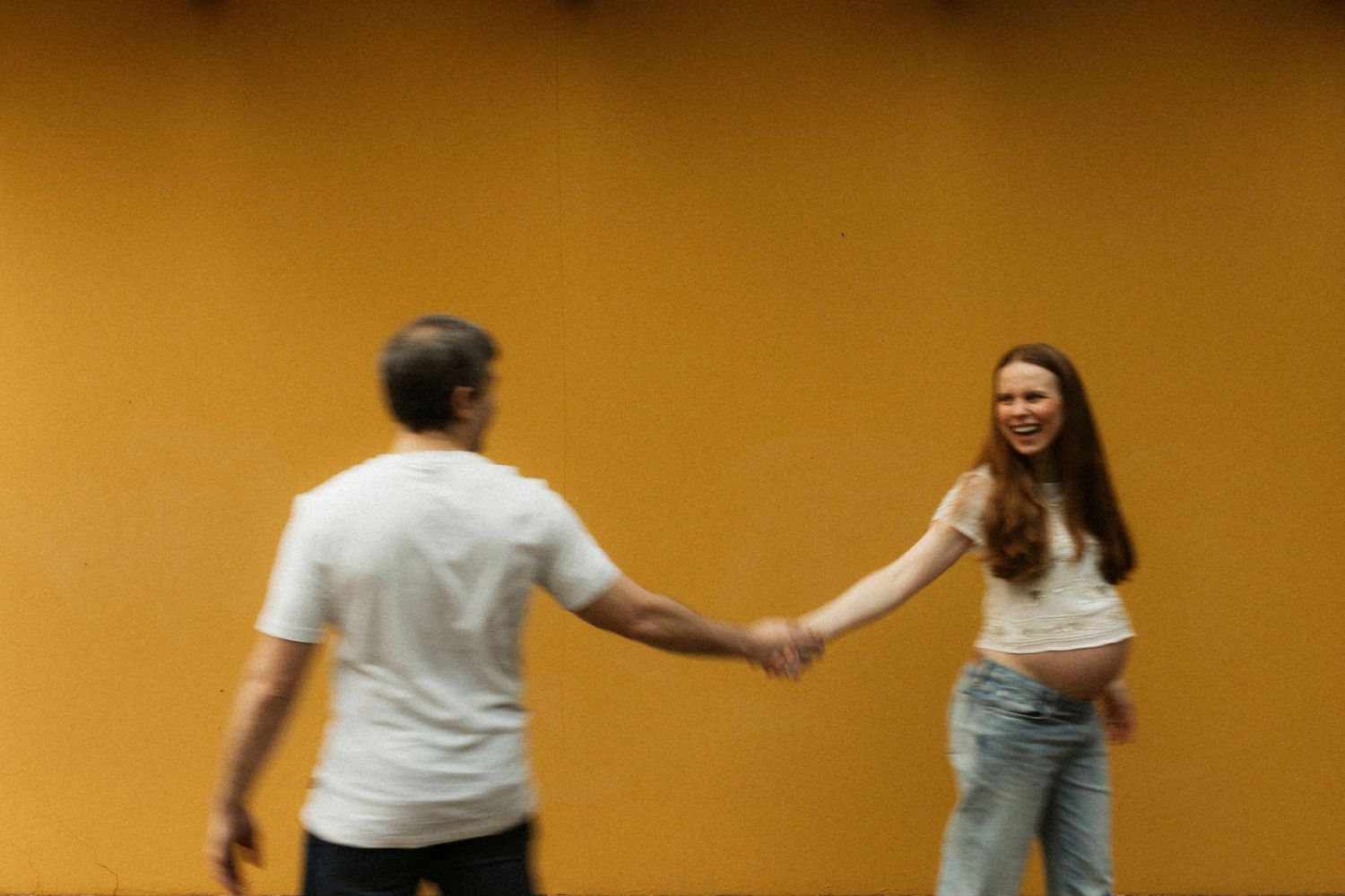 A man and woman hold hands while dancing together against a warm yellow background.