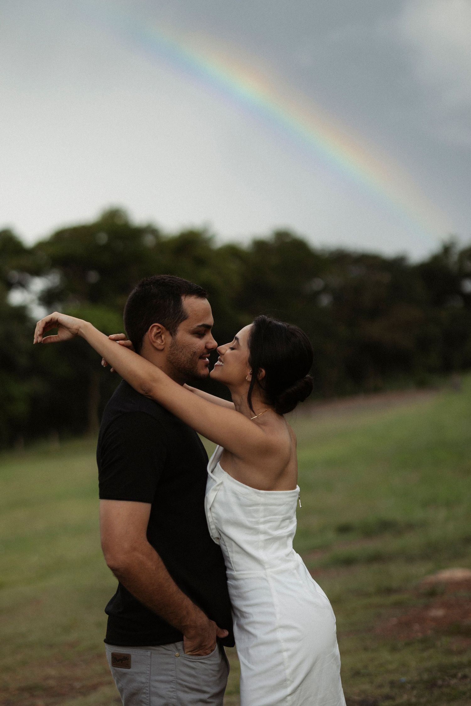 A couple shares a tender moment in a field with a rainbow visible in the cloudy sky behind them.