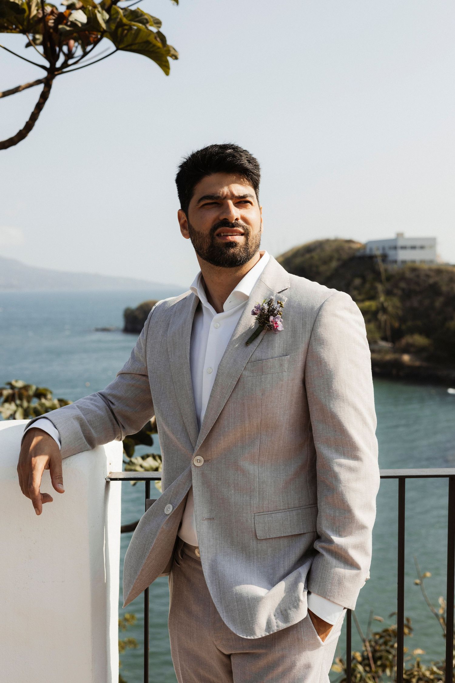 A person in a light colored suit poses against a scenic coastal backdrop on a sunny day.