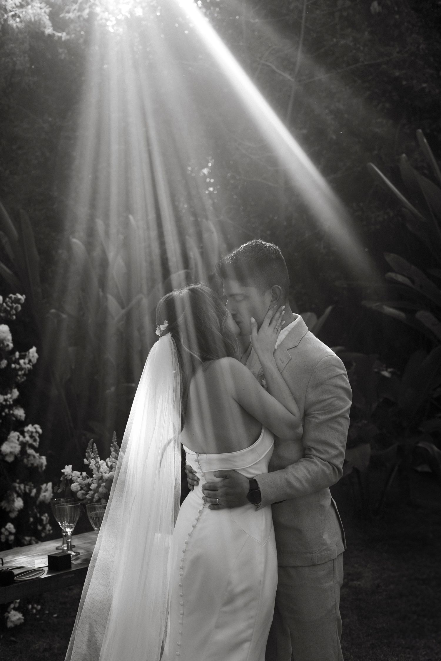 Dramatic sunbeams shine through trees onto a wedding couple in a magical forest setting.