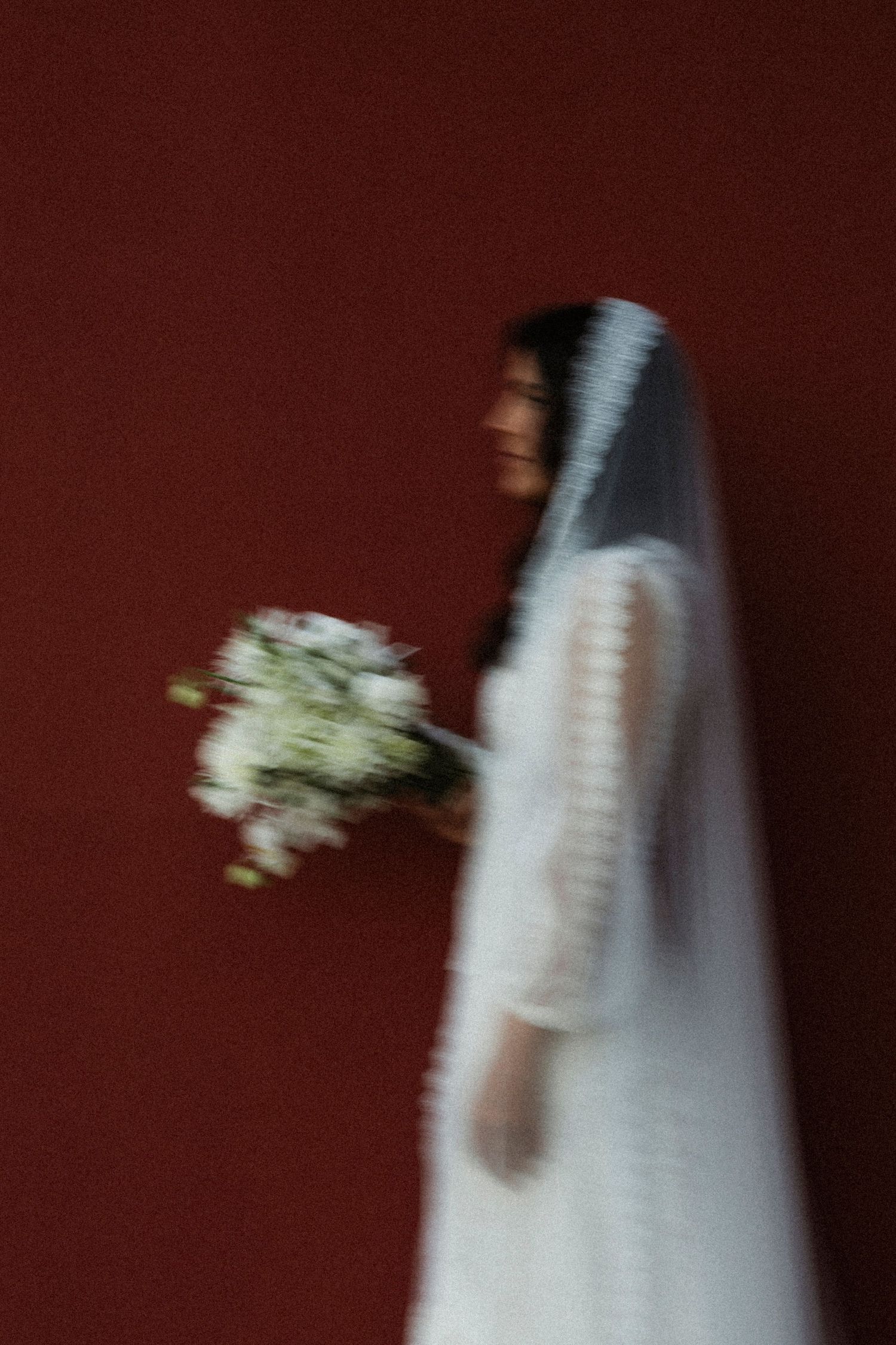 Motion-blurred image of a white wedding dress and bouquet against a dark burgundy background.