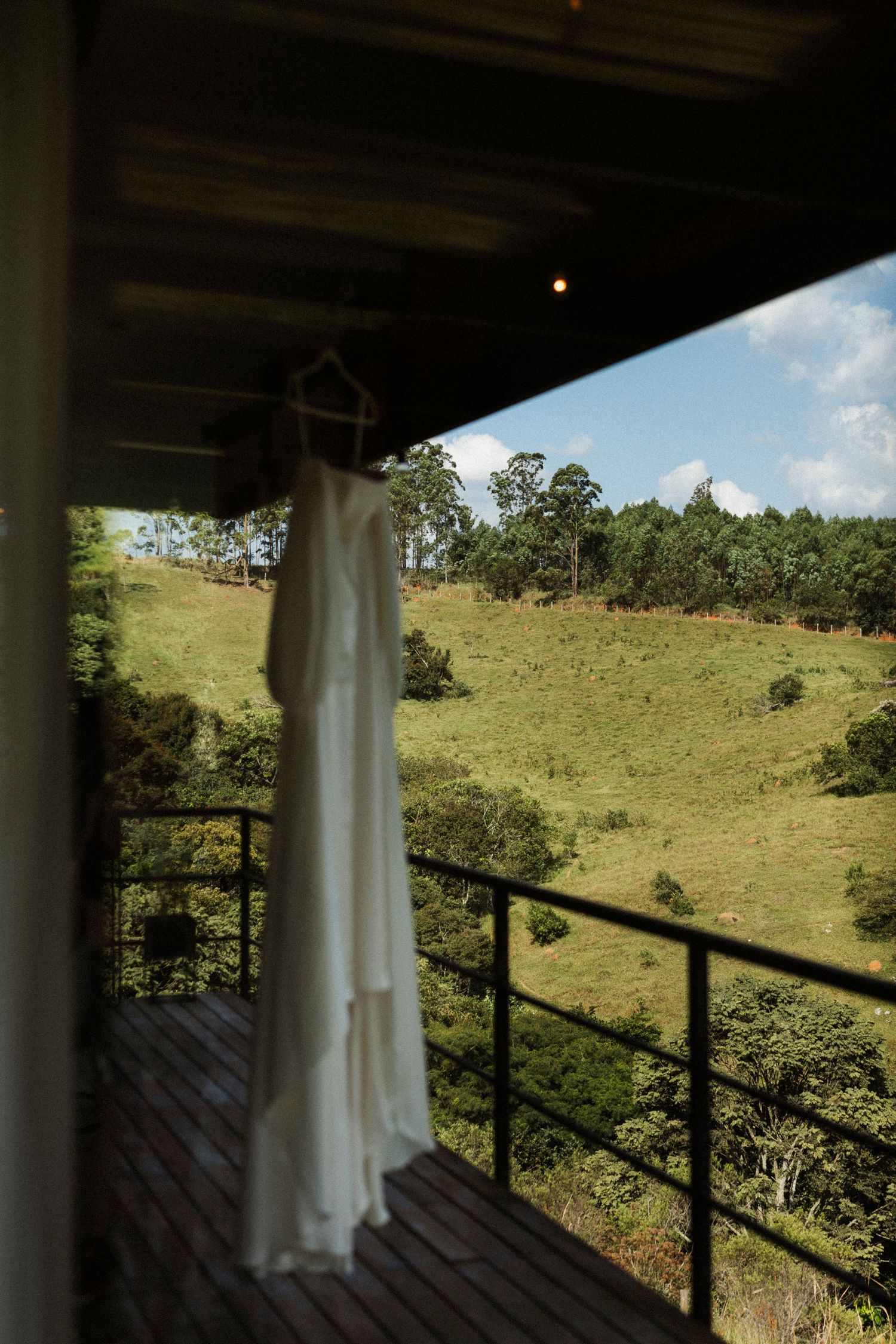 White wedding dress hanging on a balcony railing with lush green hills and blue sky in the background.
