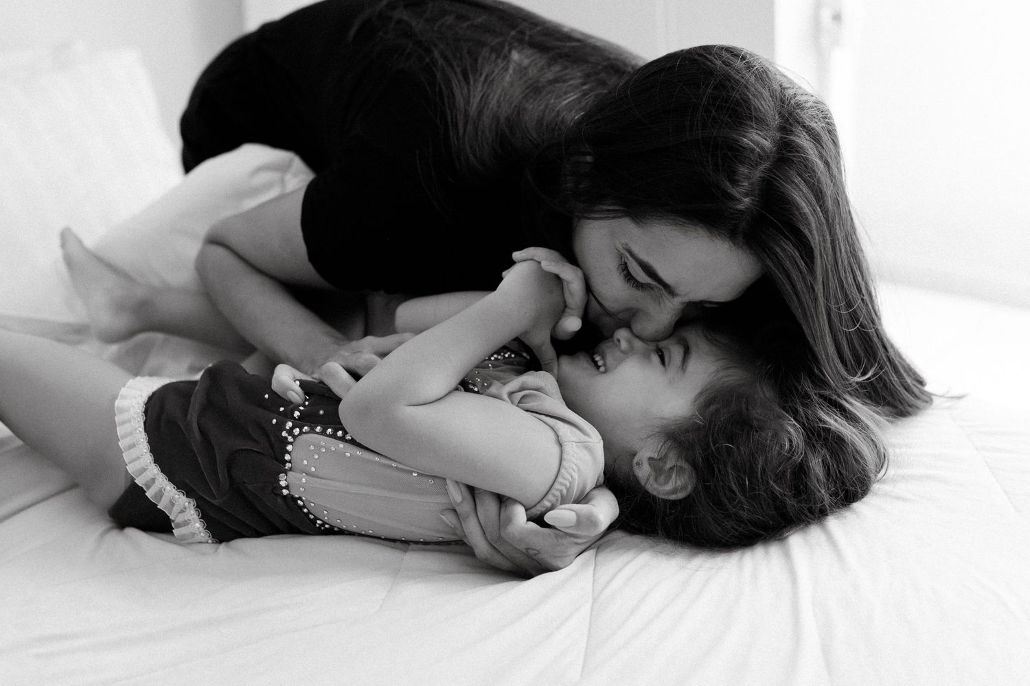Black and white photo of a loving mother kissing her young child while lying on a white bed.