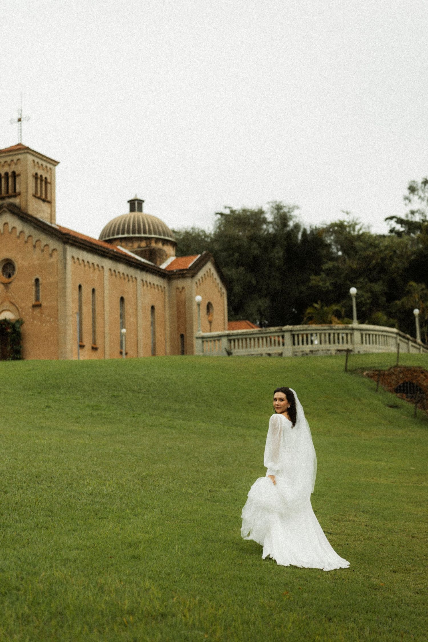 Bride in white gown and veil stands on green lawn before historic domed brick church on overcast day.