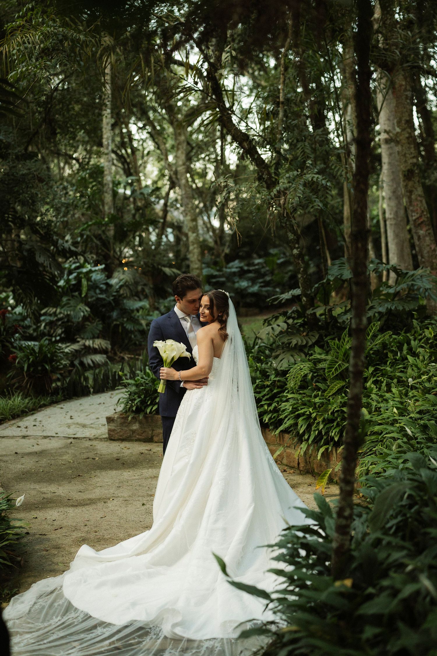 Bride in flowing white gown with veil embraces groom in navy suit amid lush green forest garden setting.