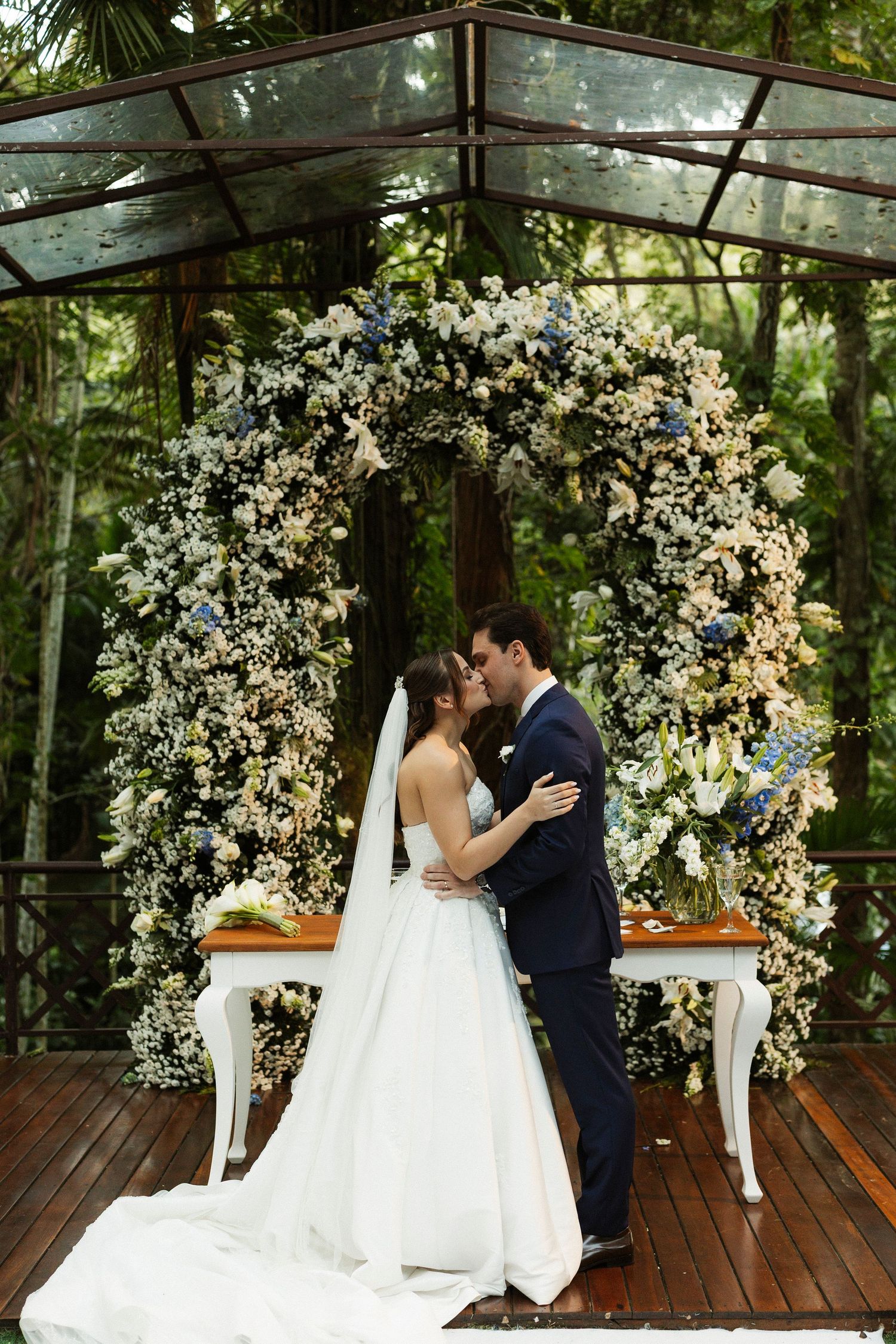 Bride and groom kissing under a floral arch with white and blue flowers at an outdoor wedding ceremony.