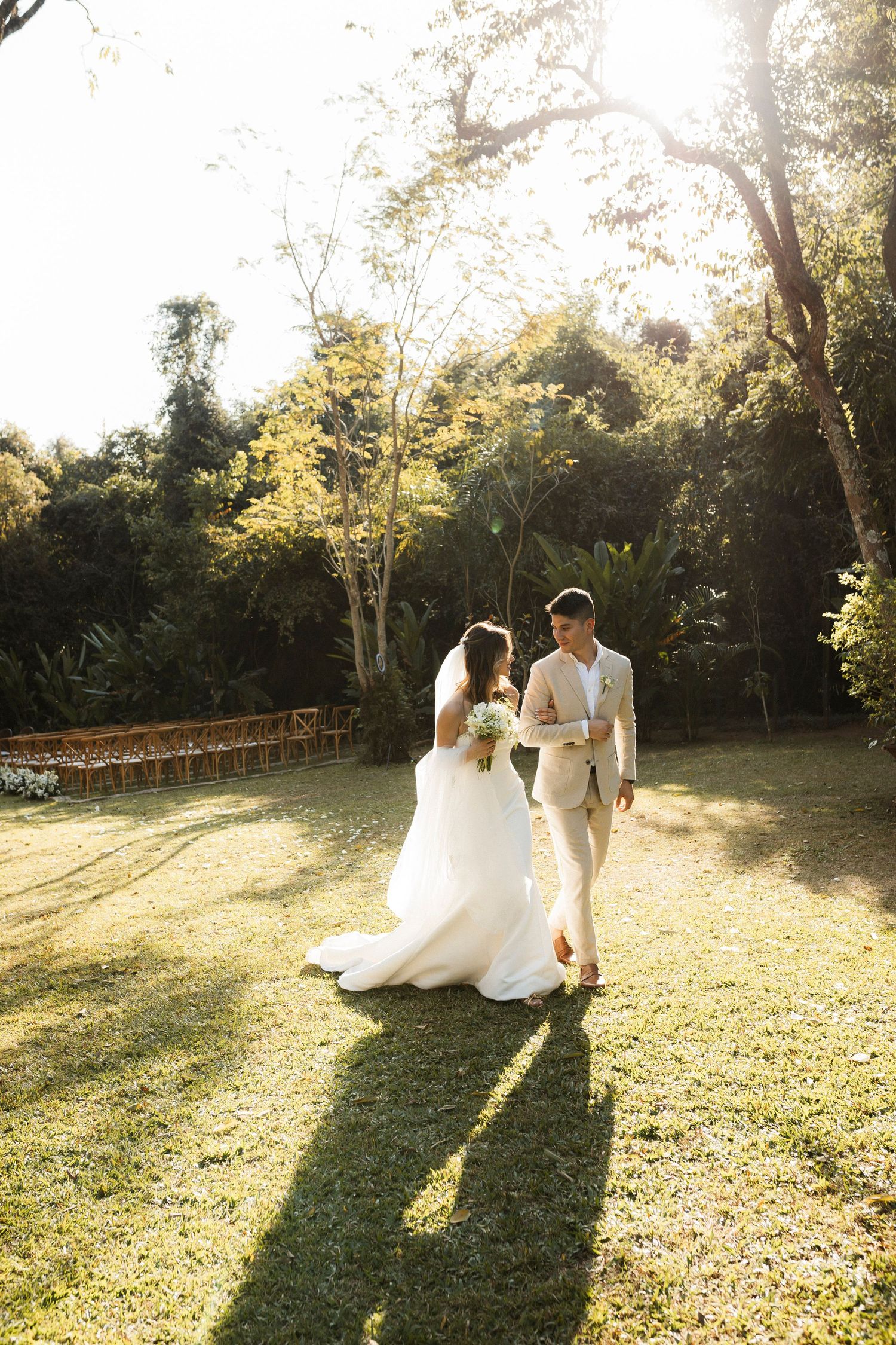 Newlyweds walk together in golden sunlight across a grassy outdoor venue.