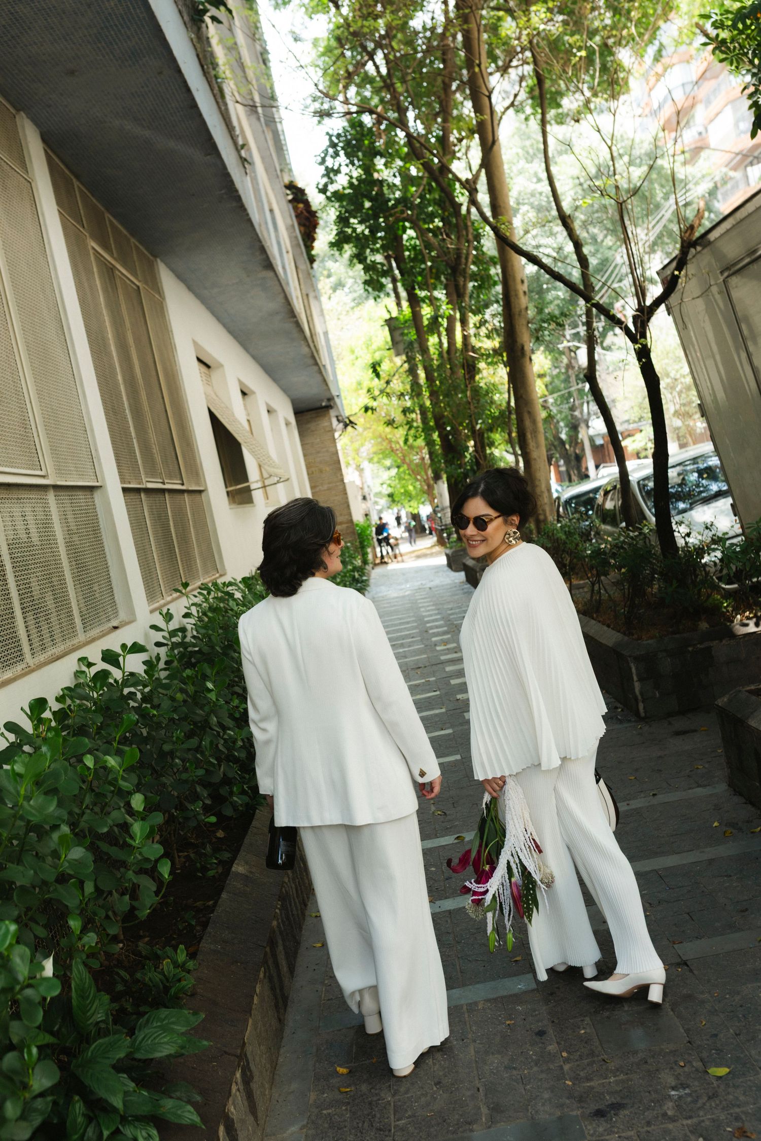 Two people in white outfits walk along a tree-lined pathway with architectural columns.
