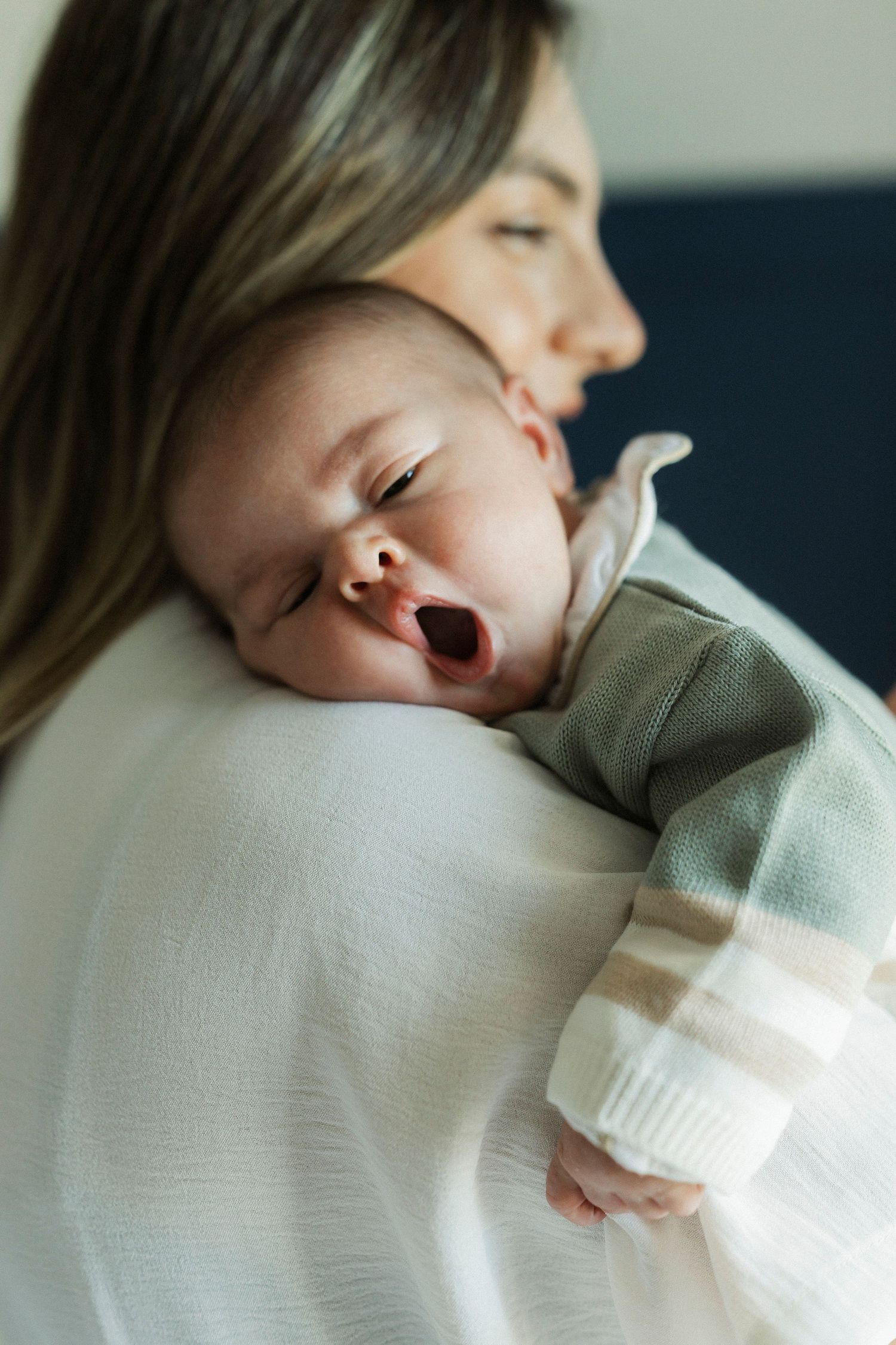 A newborn baby yawning while resting on mother's shoulder, dressed in a green striped onesie.