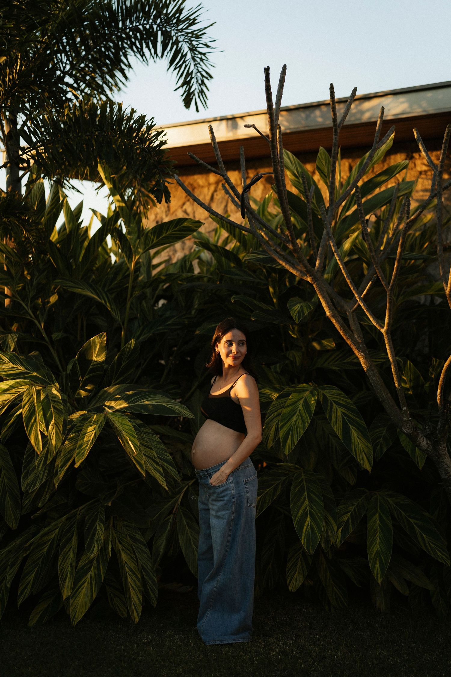 Pregnant woman in black crop top and jeans standing outdoors among tropical plants in golden hour light.