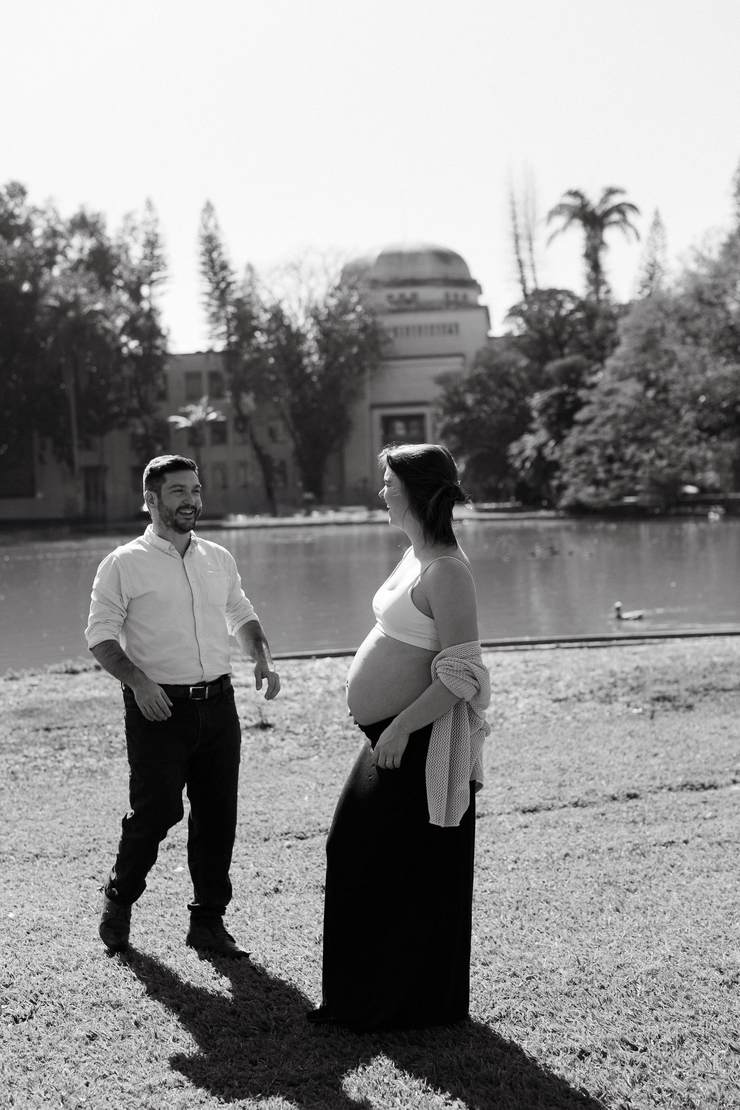 Black and white maternity photo of a couple standing by a lake in a park, woman cradling baby bump in elegant dress.