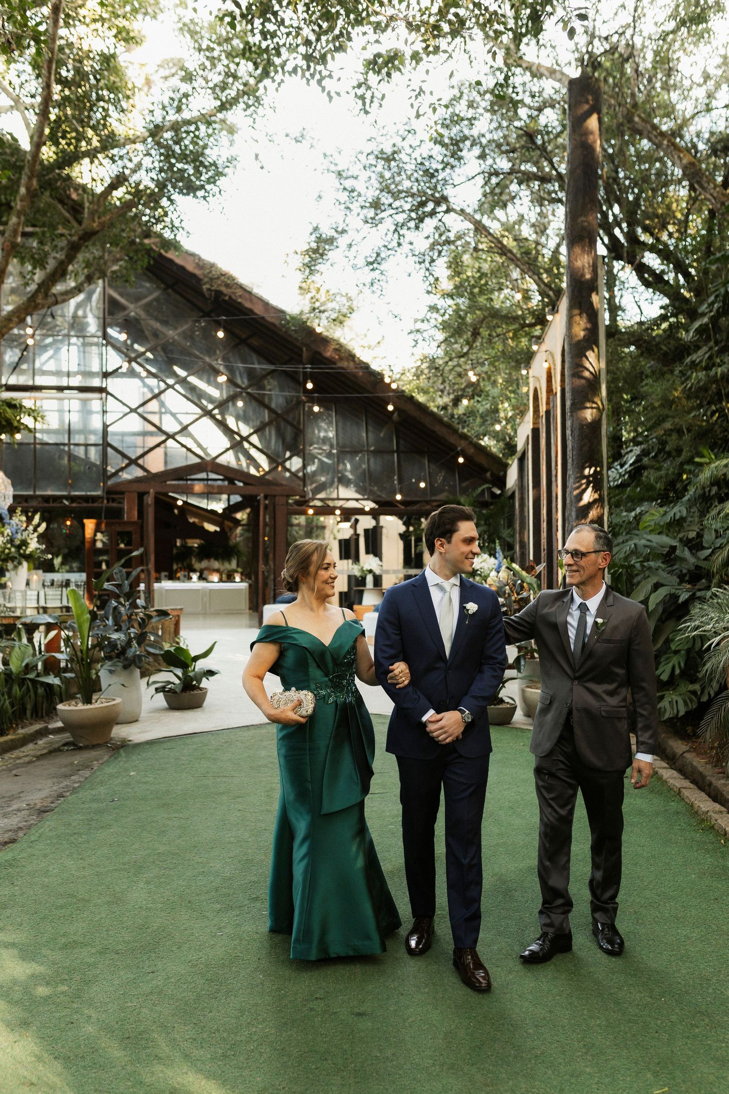 Groom in navy suit walks with parents in lush garden venue, mother in emerald gown, string lights overhead.