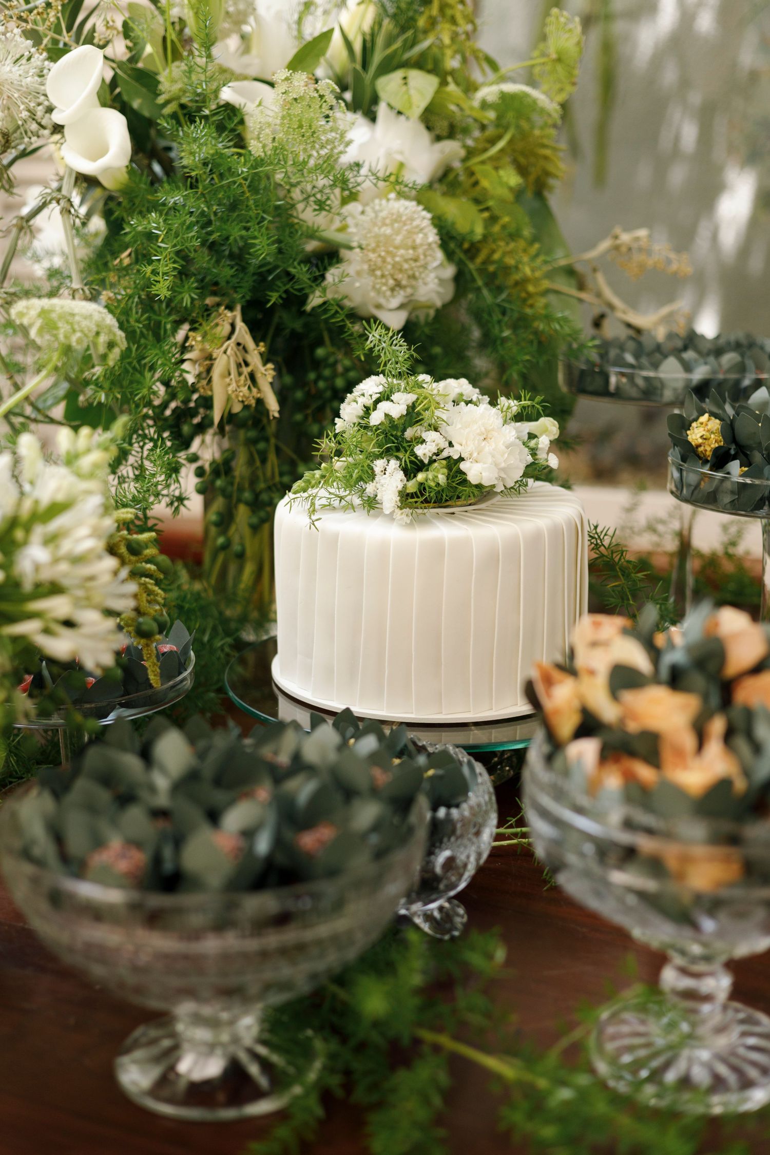 Elegant white wedding cake decorated with fresh greenery and flowers on crystal cake stands.