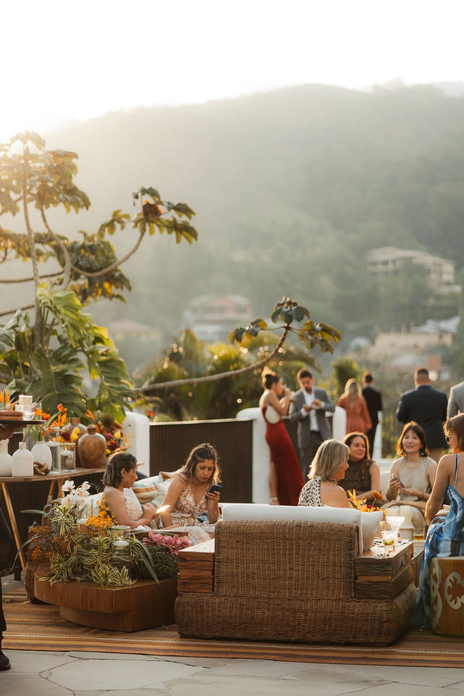 Wedding guests relax on wicker furniture at an outdoor reception with mountain views at sunset.