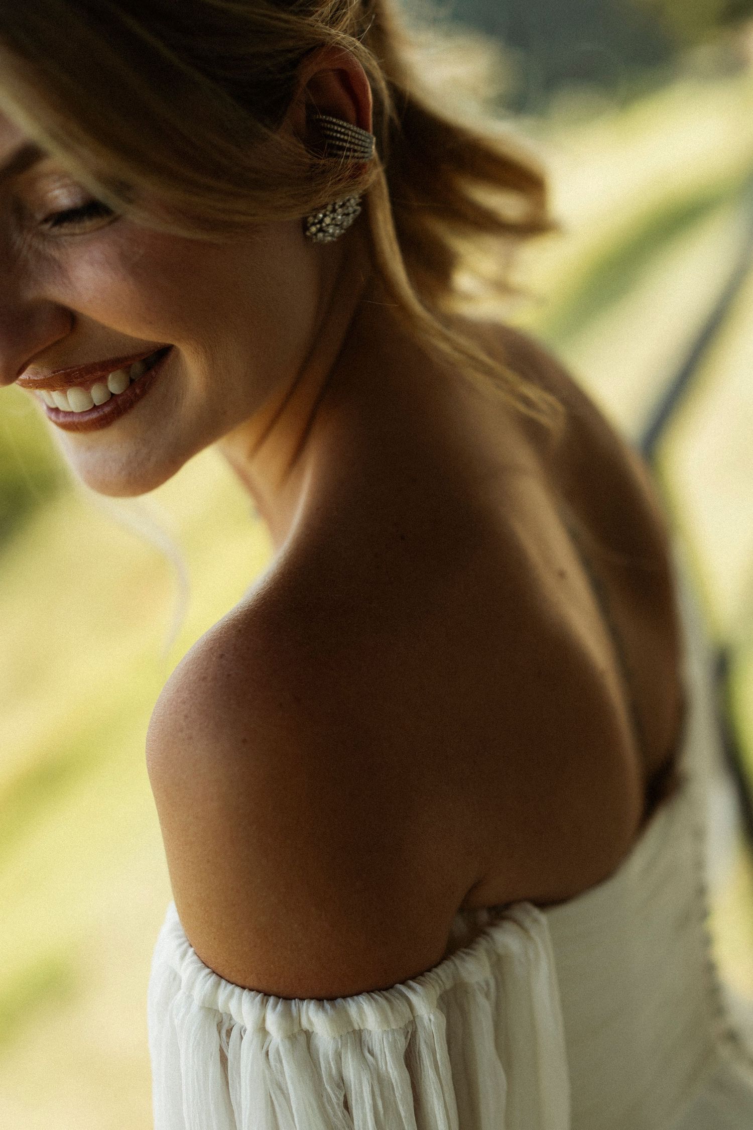 Smiling woman in white off-shoulder dress with elegant crystal earring, photographed outdoors in warm natural light.