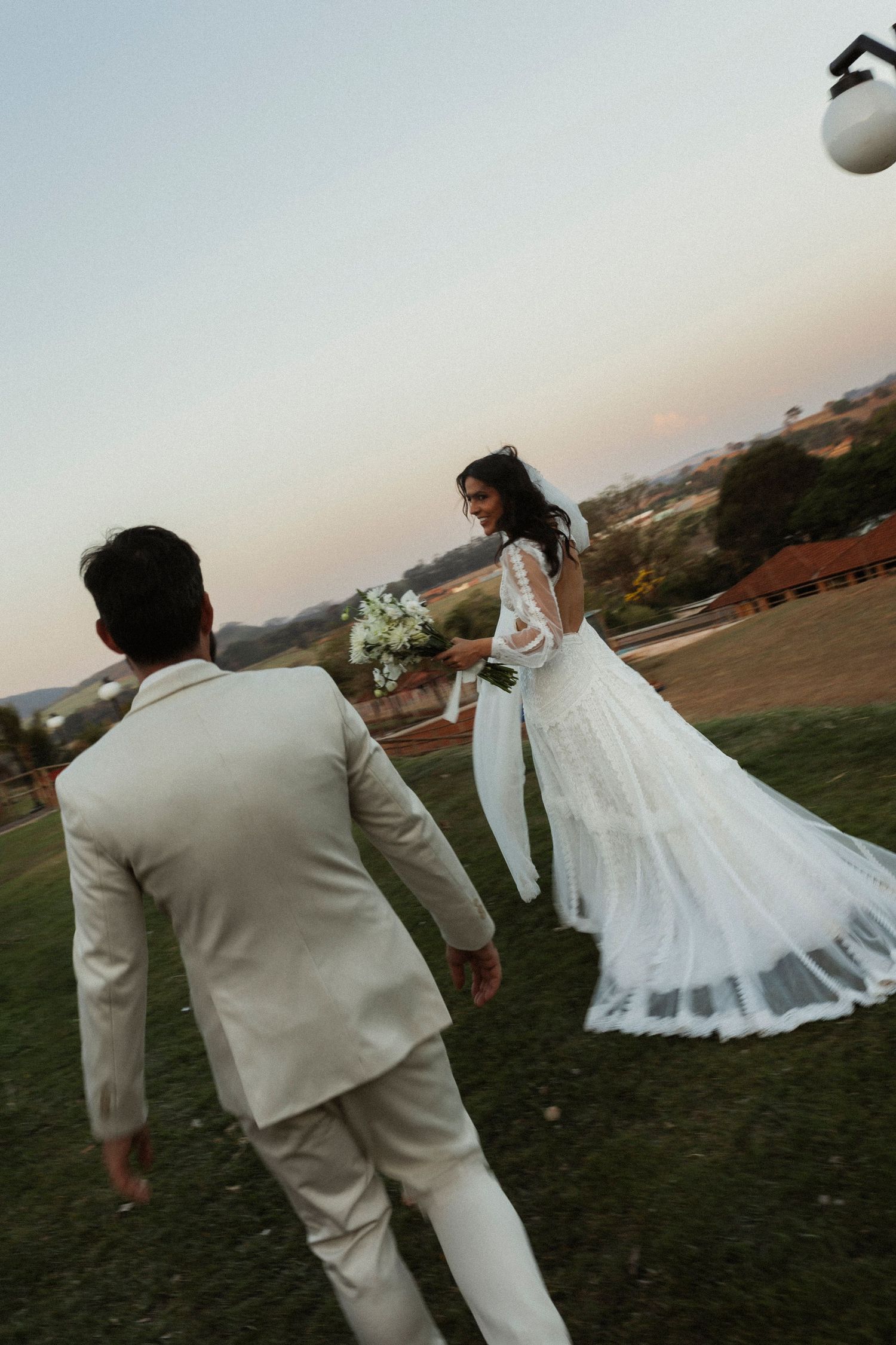Newlyweds in white attire walk hand in hand across grassy field at dusk.