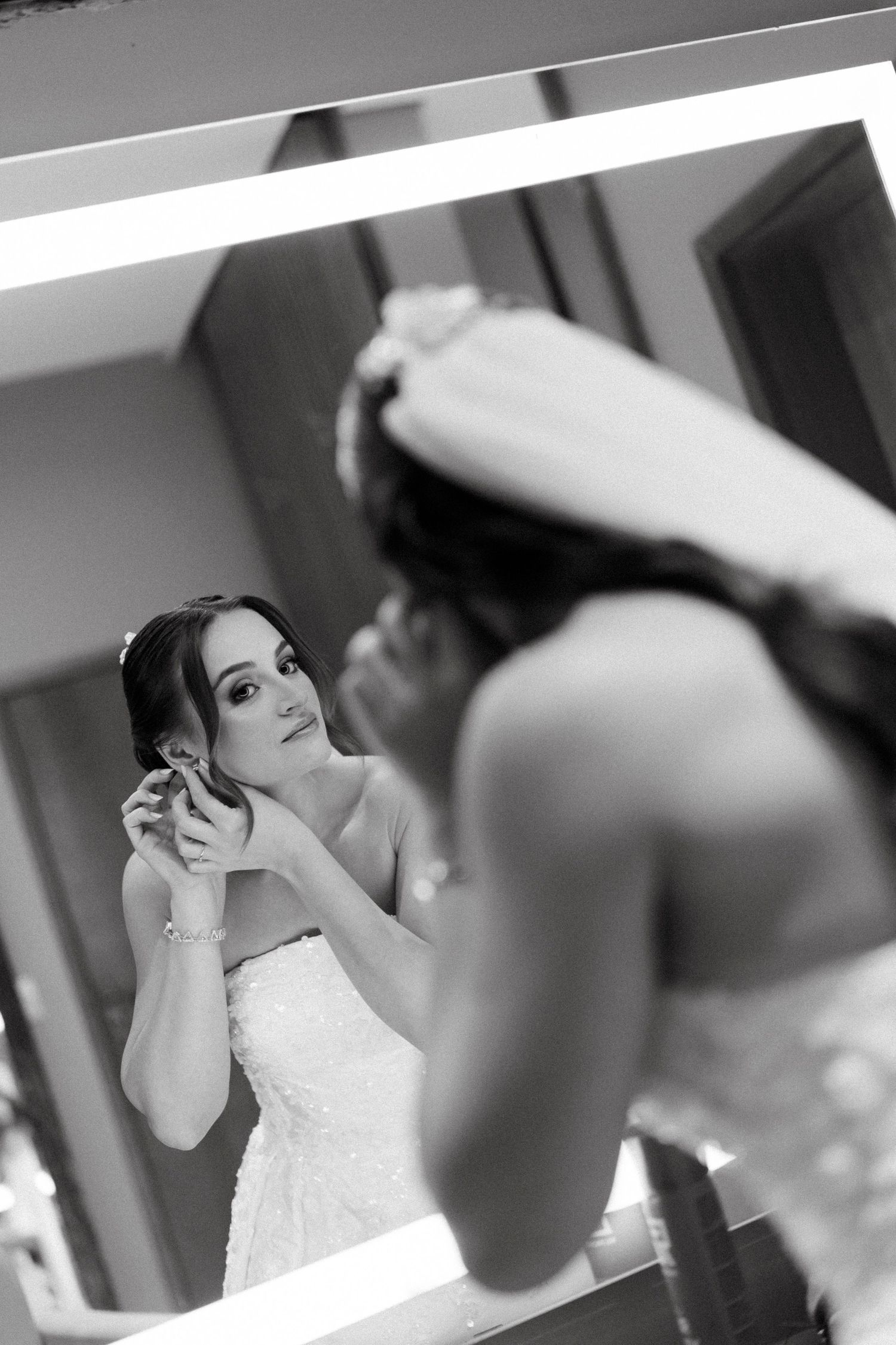 Bride in strapless wedding dress putting on earrings while looking in a lit mirror, black and white photo.