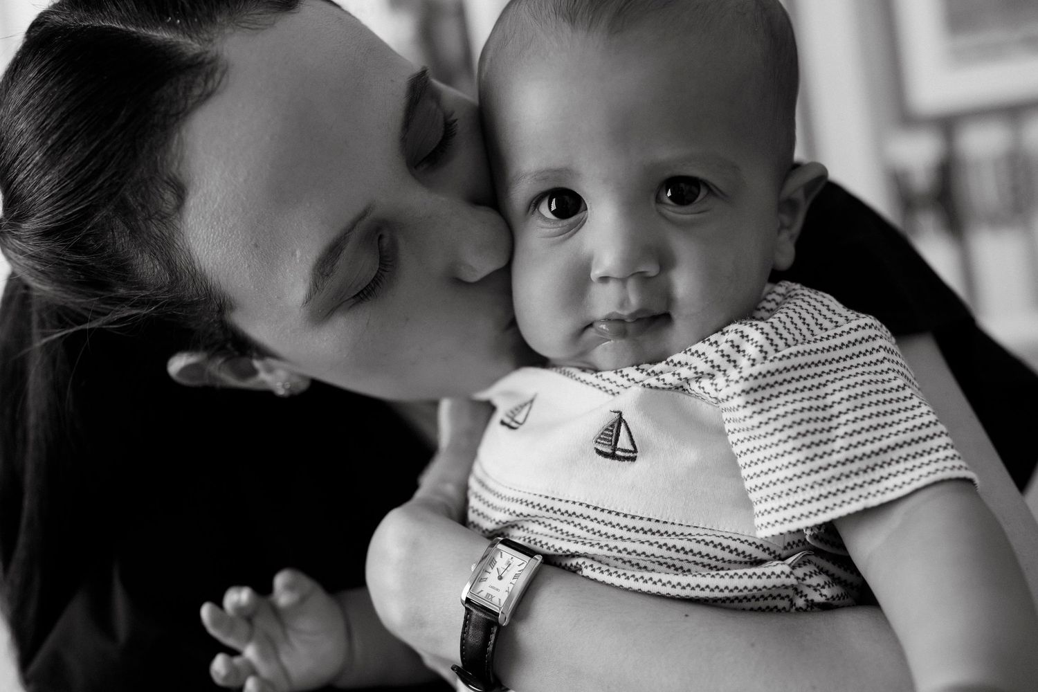 Black and white photo of a mother kissing her baby boy wearing a striped outfit with a sailboat embroidery.