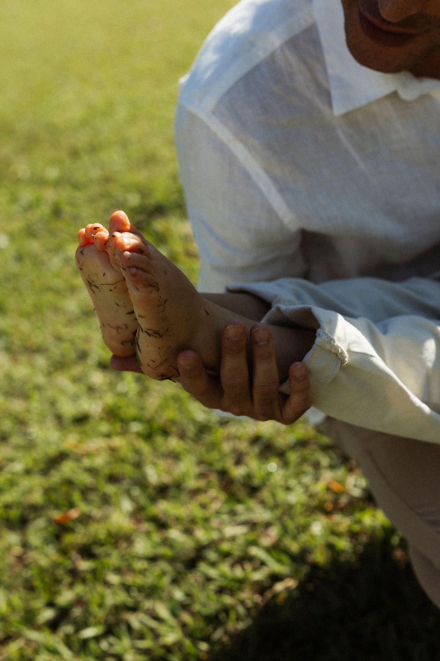 Person in white clothing holding feet with henna designs while sitting on green grass outdoors.