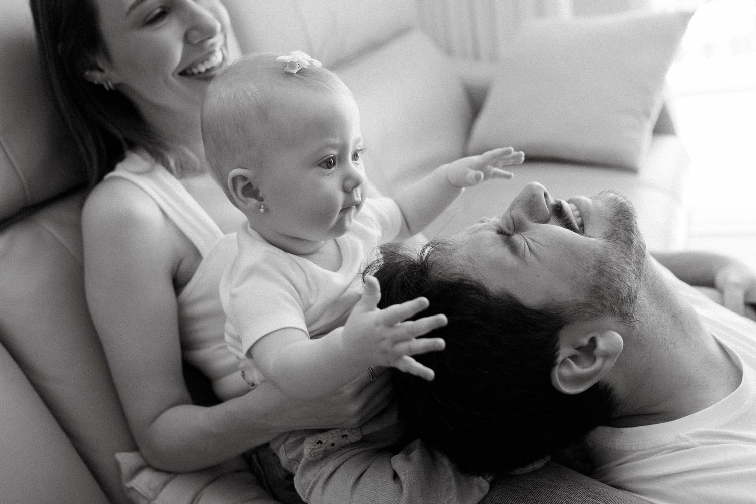 Black and white photo of happy parents smiling and playing with their baby on a couch at home.