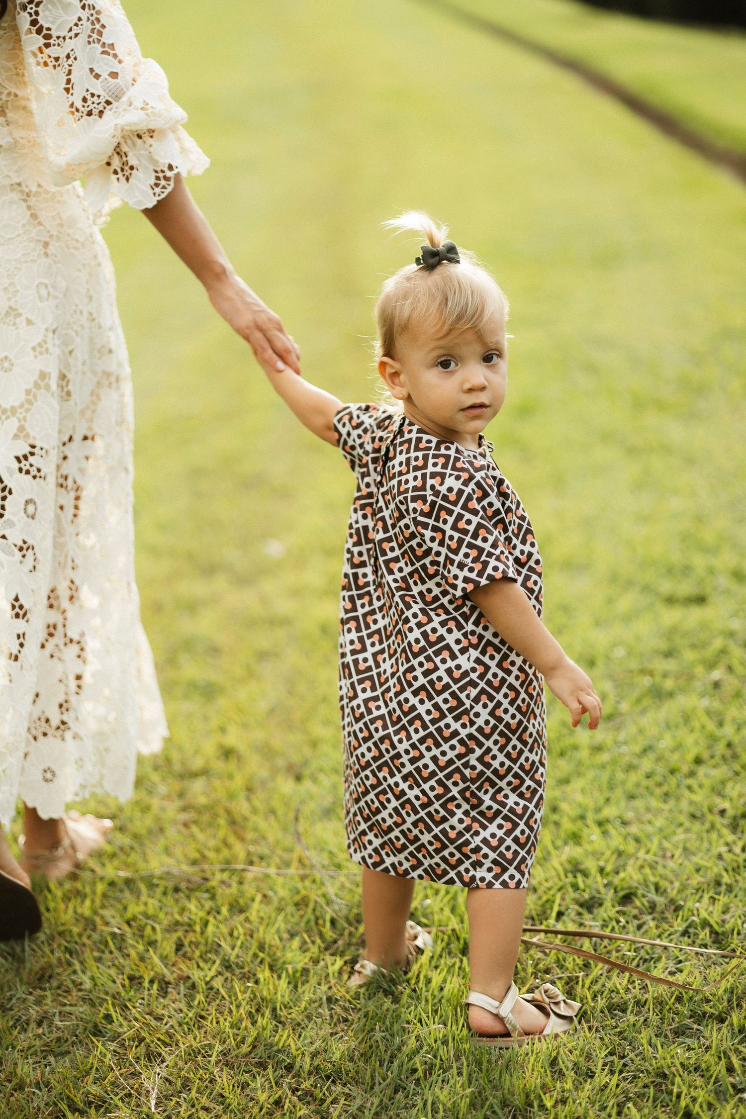 Toddler girl in patterned dress holds mother's hand while walking on green grass in warm sunlight.