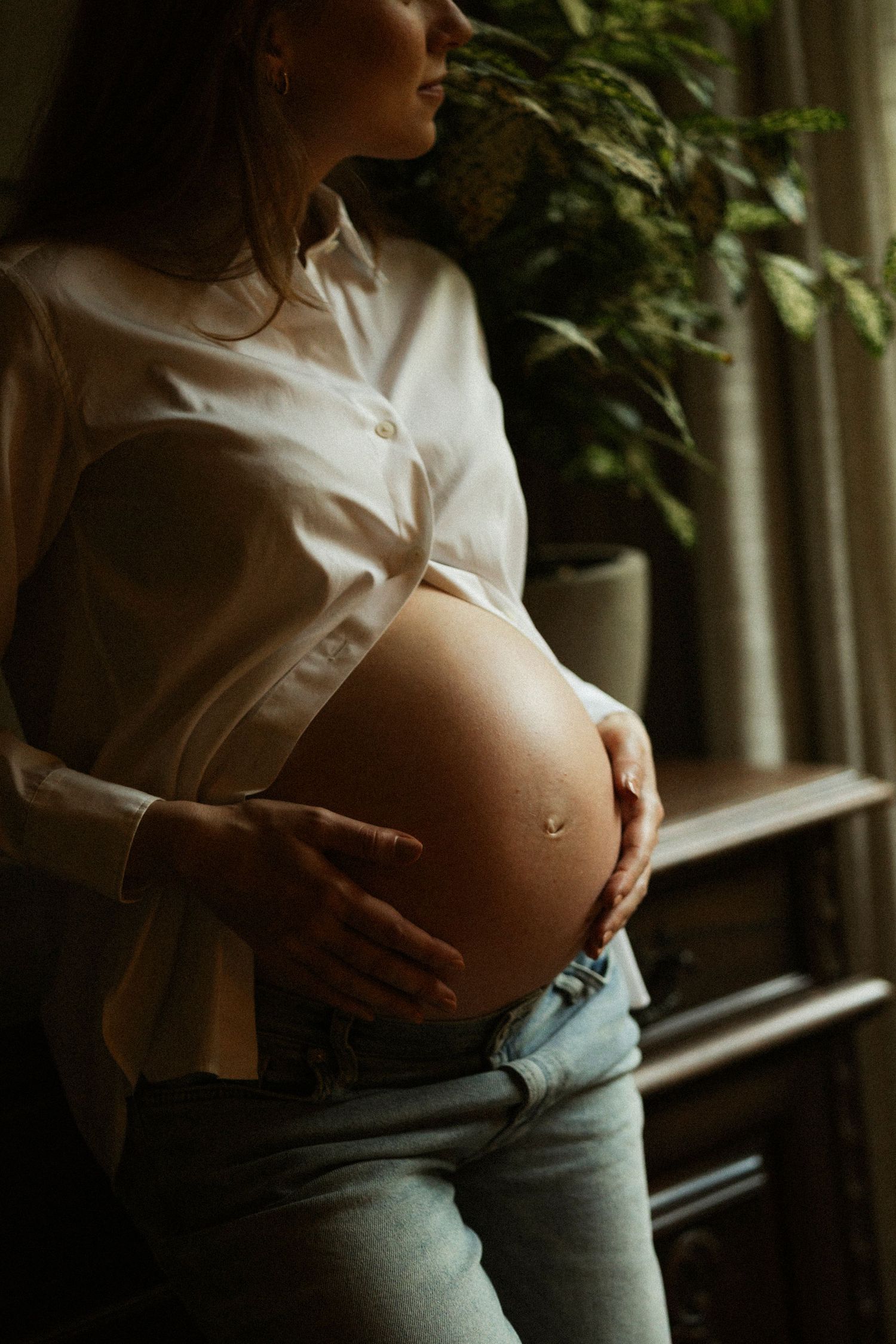 Pregnant woman in white shirt cradling baby bump by window with natural light and plants in background.