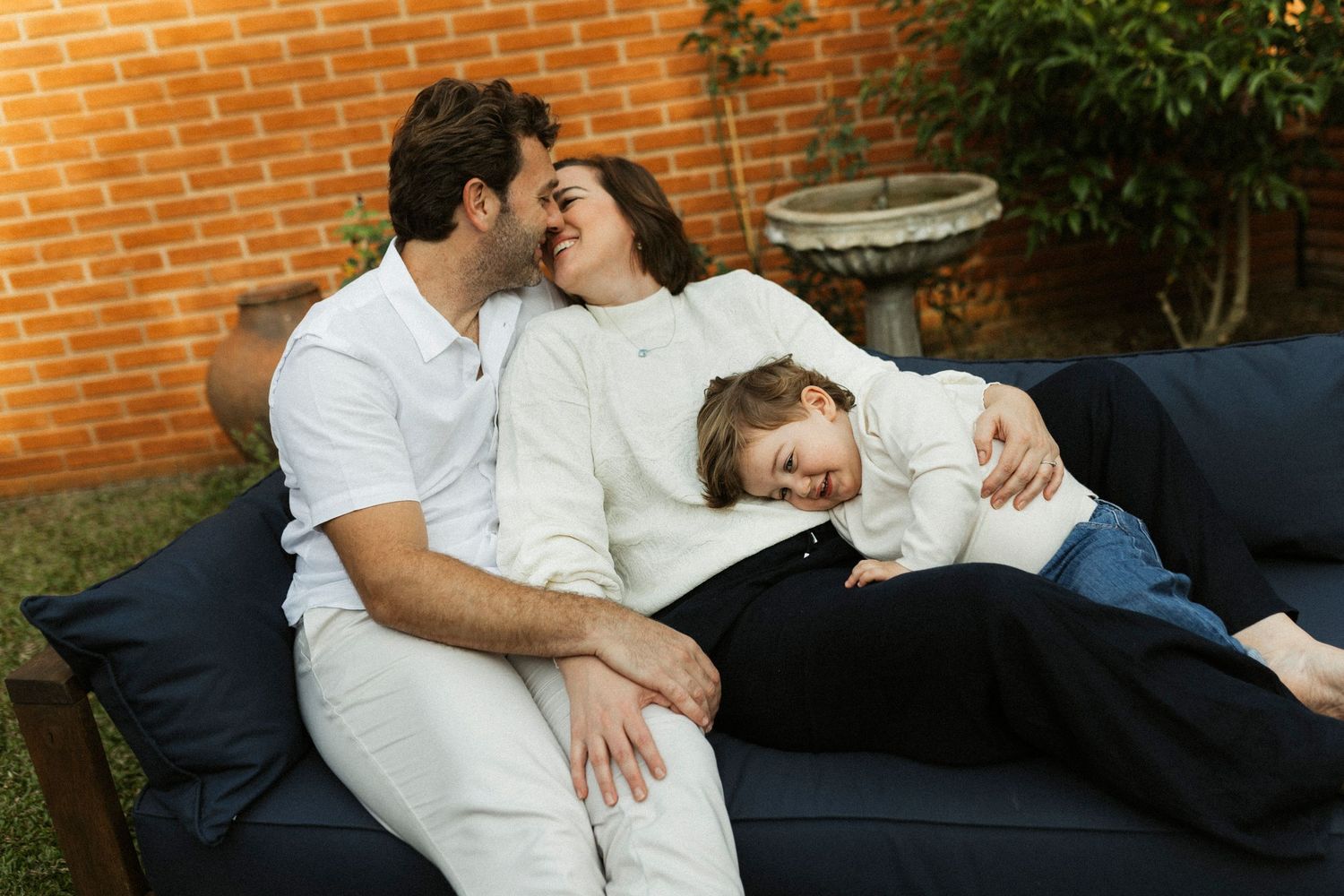 Family of three relaxing together on a navy outdoor sofa, sharing a warm embrace in a cozy backyard setting.