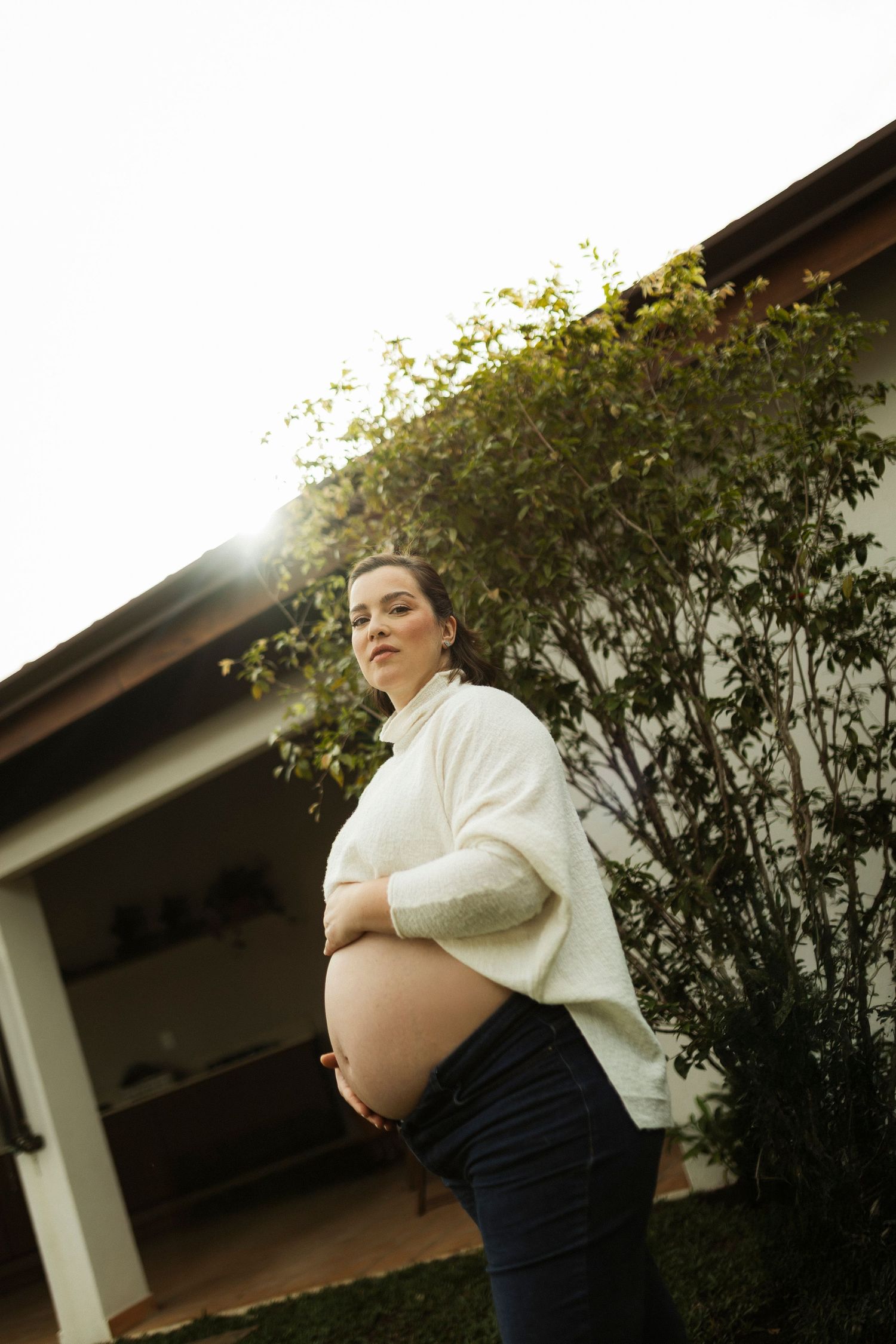 Pregnant woman in white sweater cradles bare belly outdoors, standing in front of a house with green shrubs.