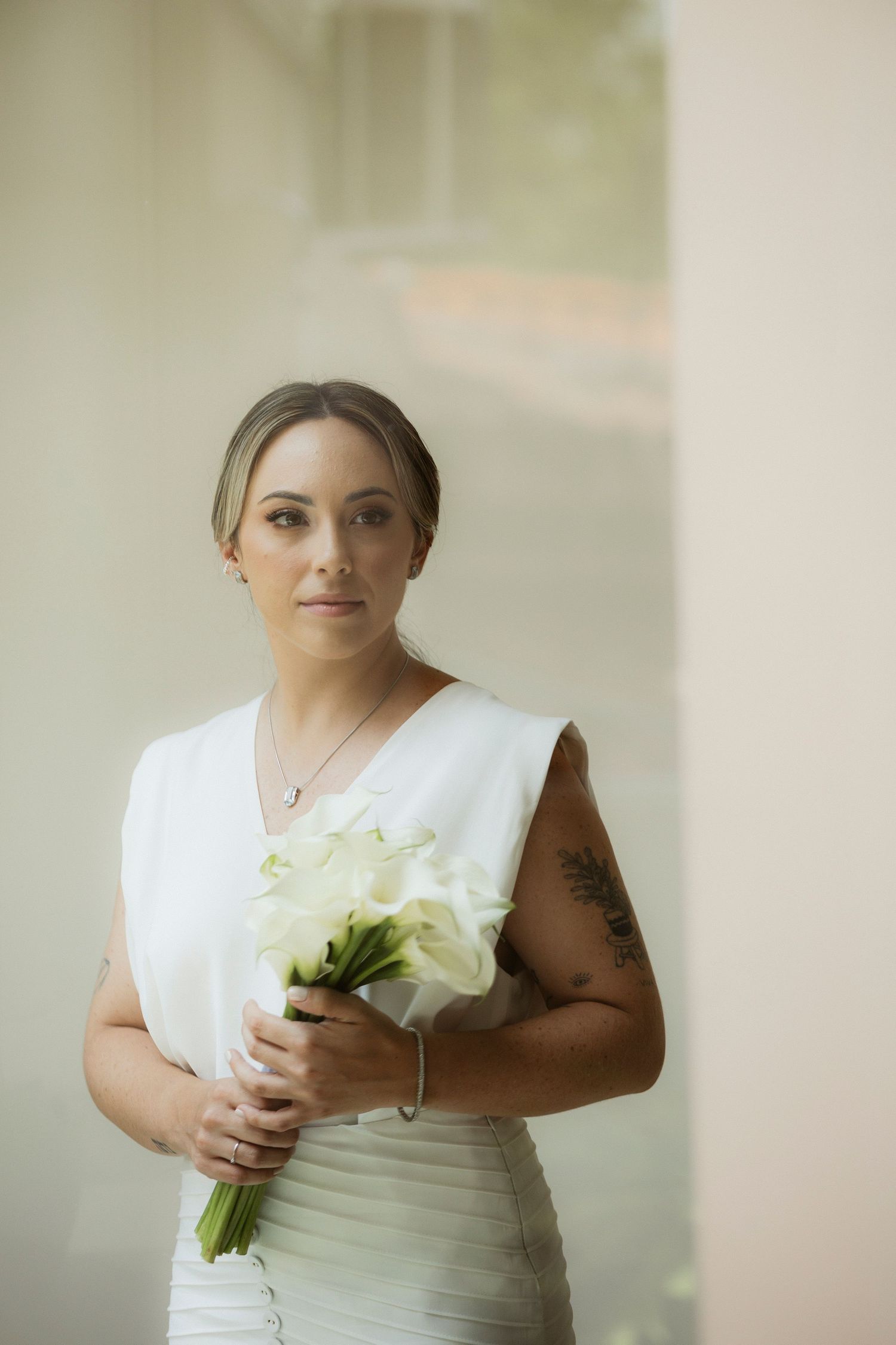 Elegant bride in sleeveless white dress holds bouquet of white calla lilies against soft background.