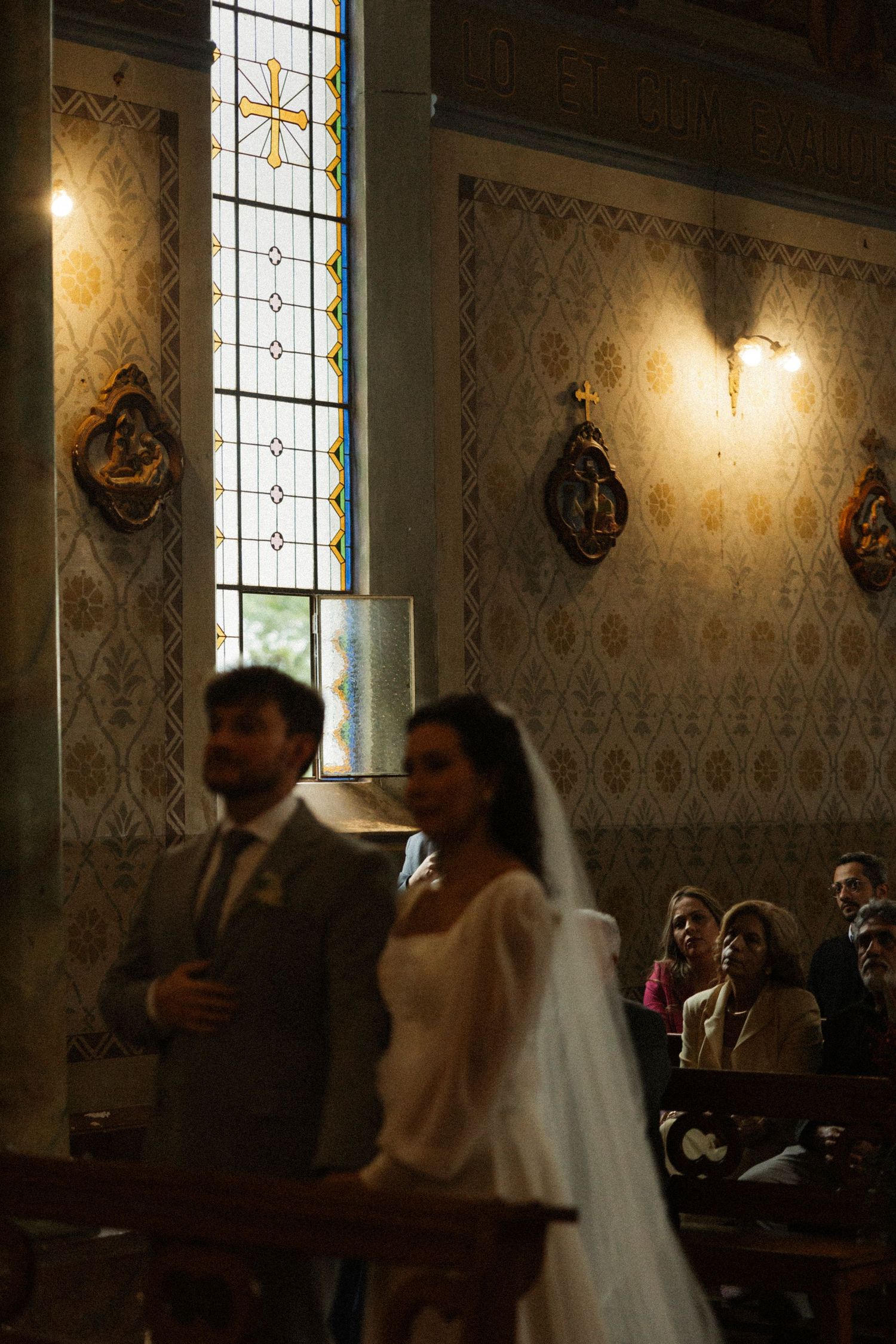Bride in white veil and groom stand at church altar during wedding ceremony with stained glass windows.