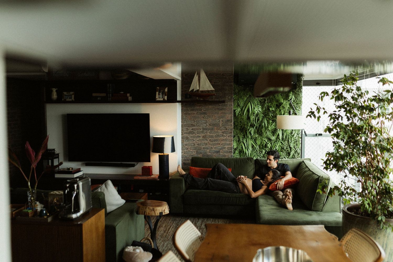 Couple relaxing on a green sofa in a modern living room with TV, brick wall, and lush vertical garden.