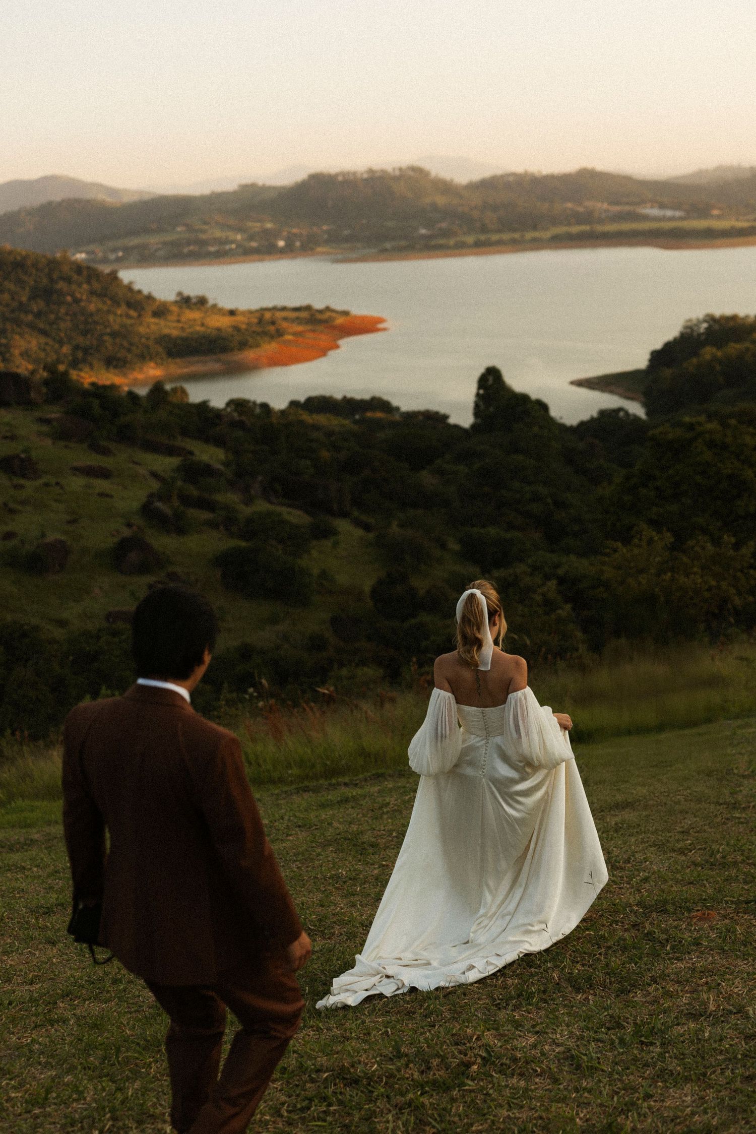 Bride in white gown and groom in brown suit overlook scenic lake and green hills at golden hour.
