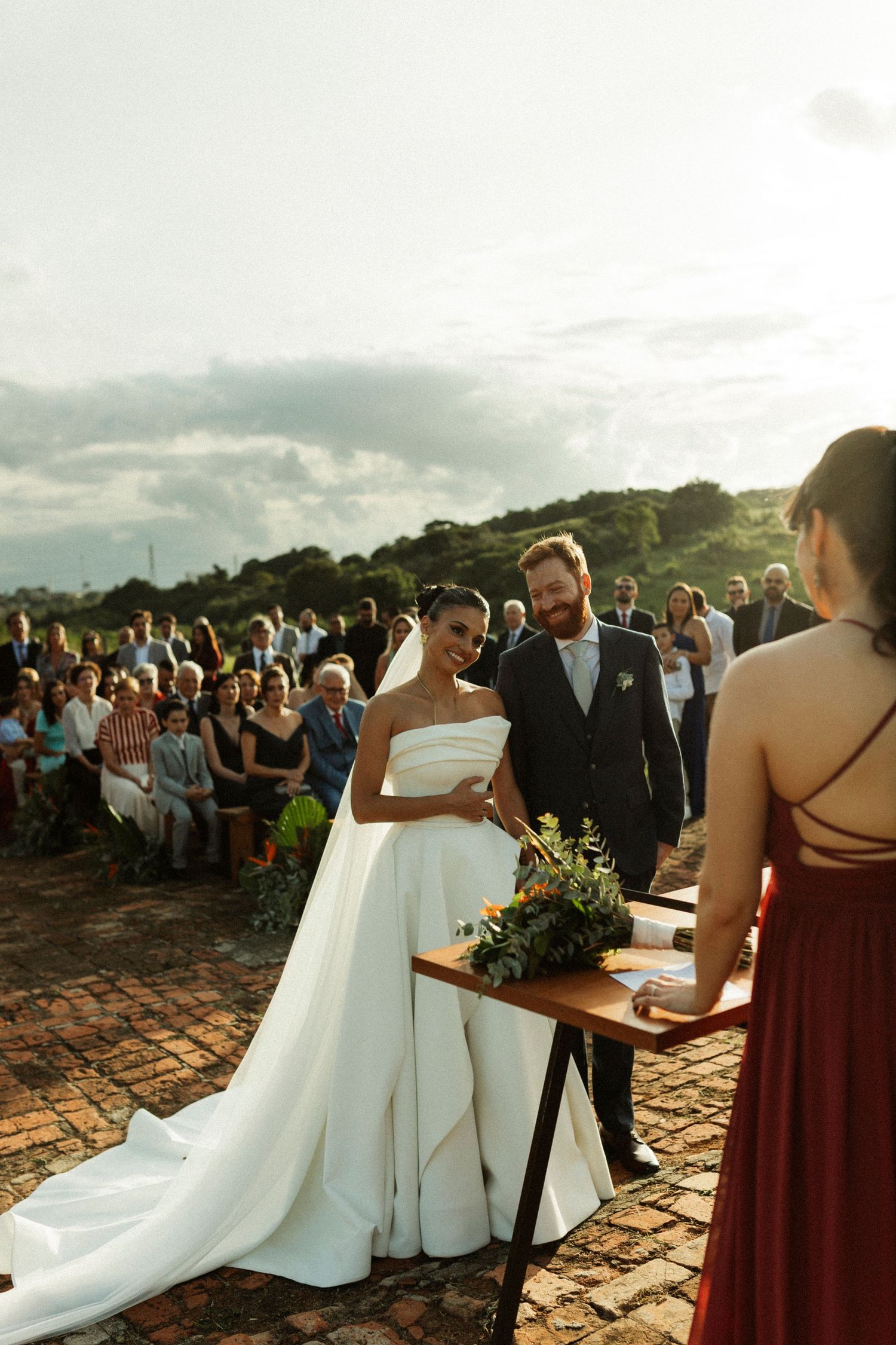 Bride and groom smile during outdoor wedding ceremony at sunset with guests seated behind them.