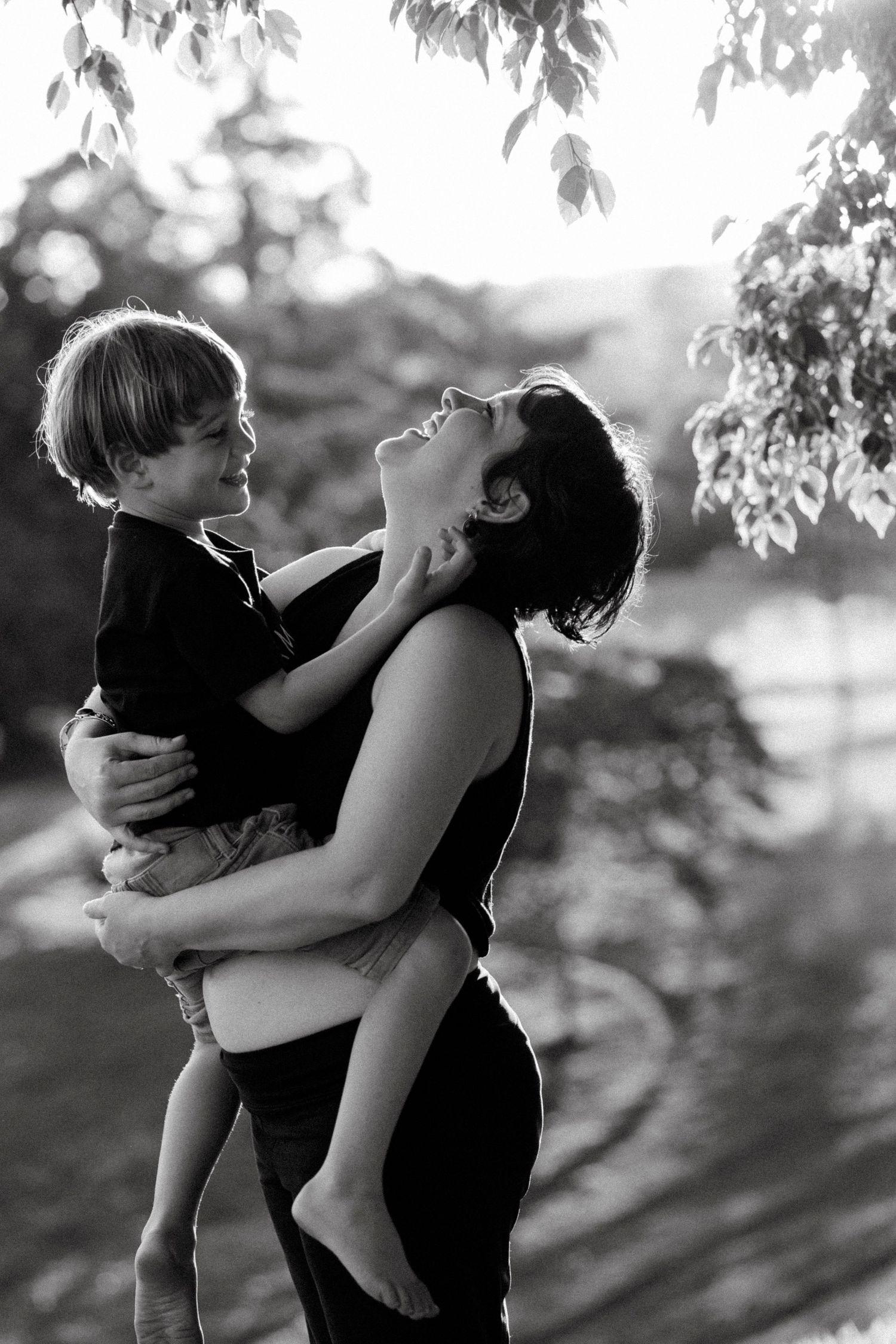 Black and white photo of a laughing mother holding her young child outdoors beneath leafy trees.
