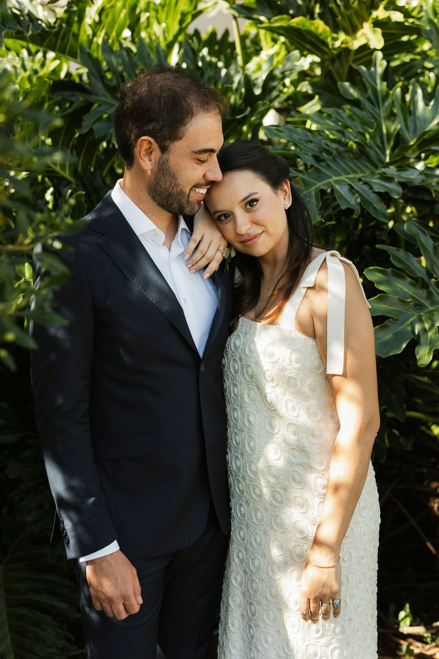 A couple in formal attire embraces outdoors, woman in white dress leans on man in dark suit amid lush tropical greenery.