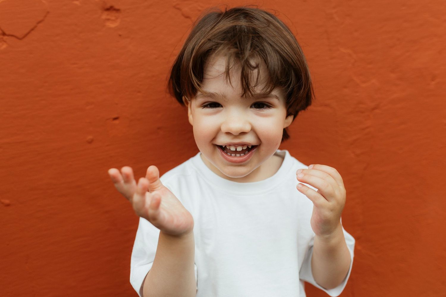 Smiling child in white shirt against orange wall making playful expressions.