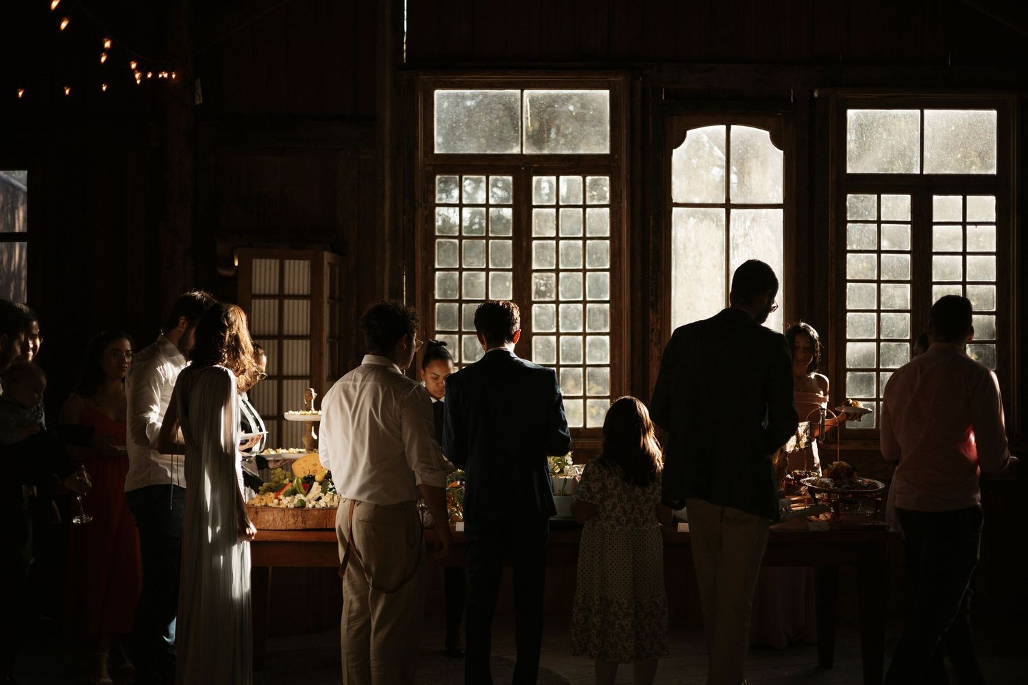 Wedding guests mingle in an elegant indoor reception venue with warm lighting and large windows.