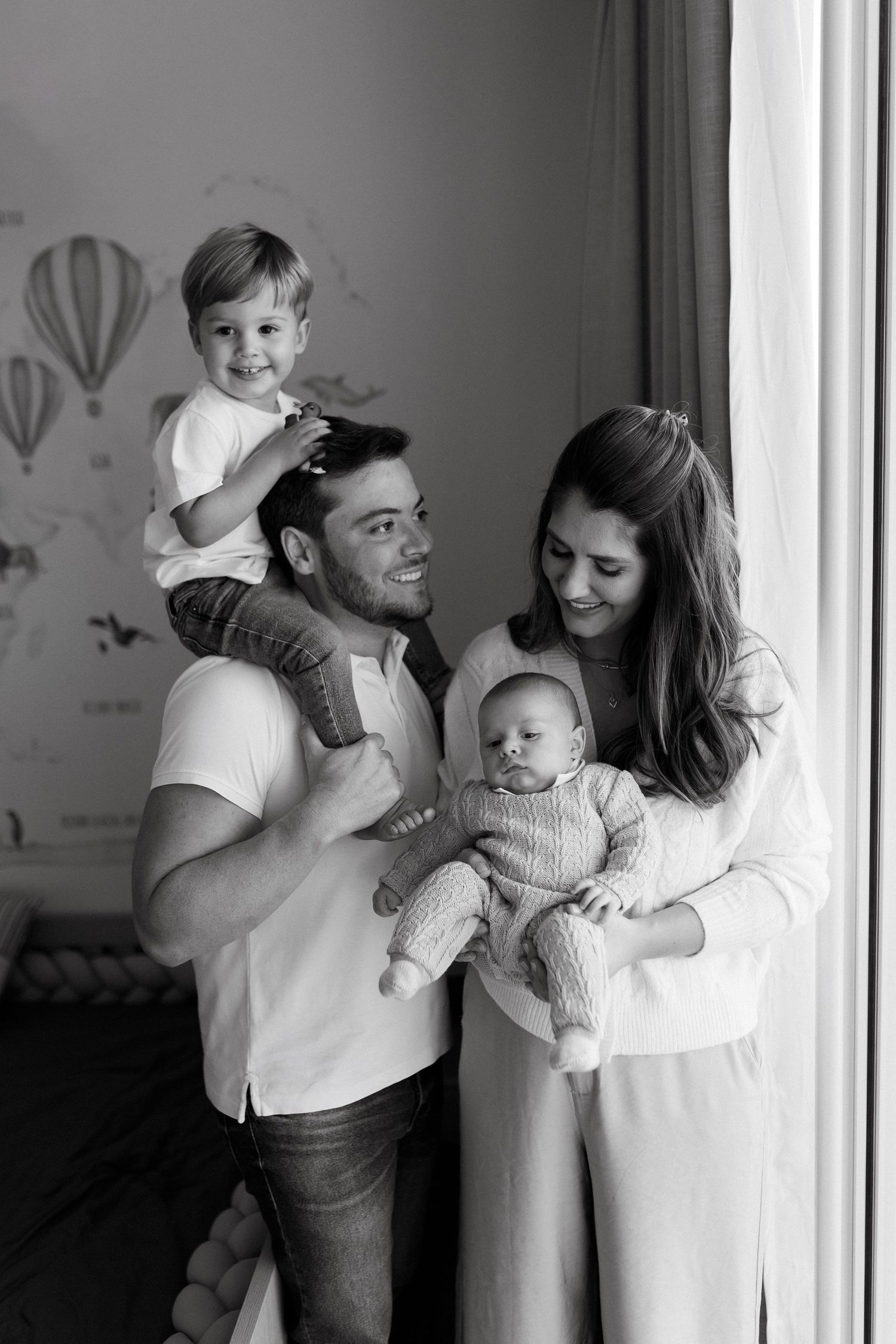 Black and white family portrait with parents holding newborn baby and toddler son near window.