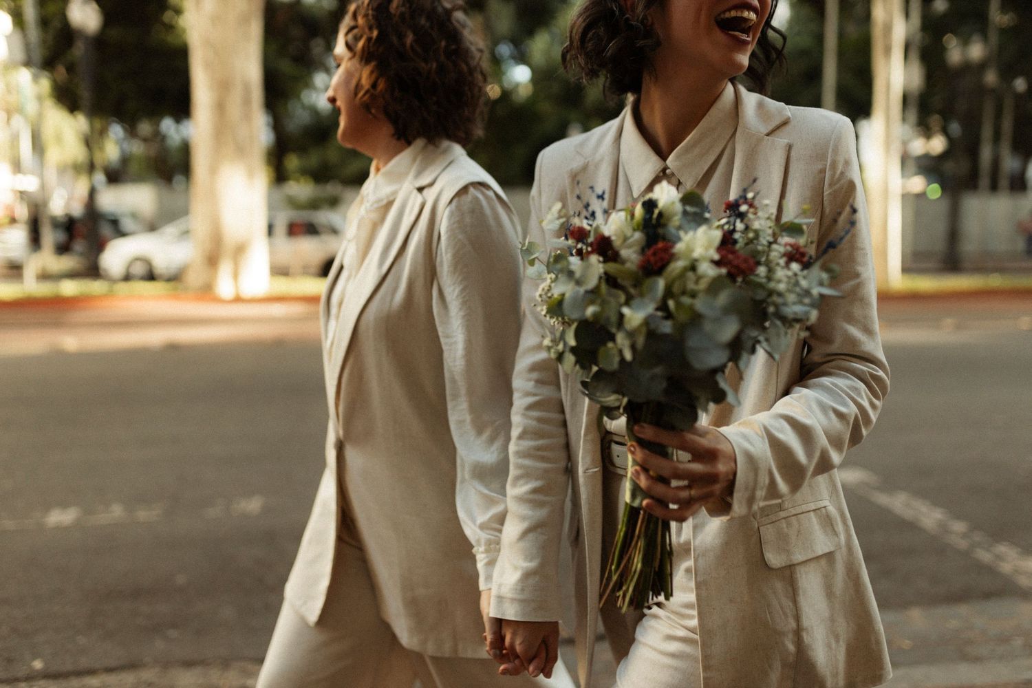 Two people in white suits walking together outdoors, one holding a beautiful floral bouquet.
