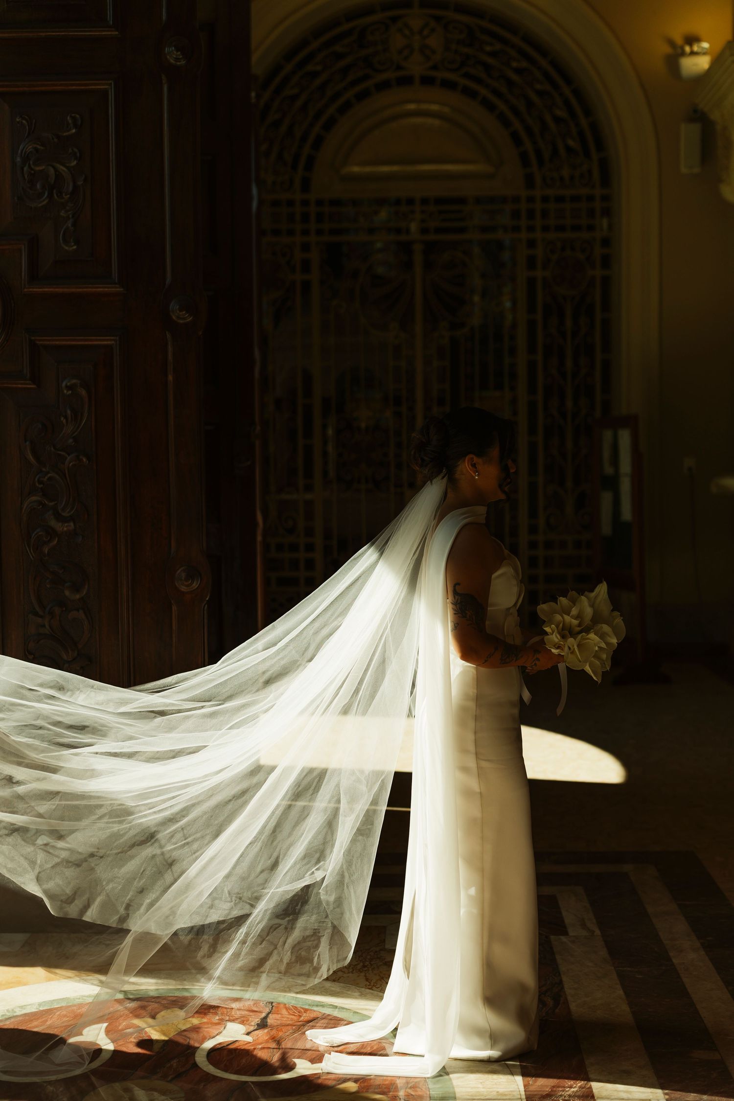 Bride in white gown with flowing cathedral veil standing in dramatic light at ornate church entrance holding bouquet.