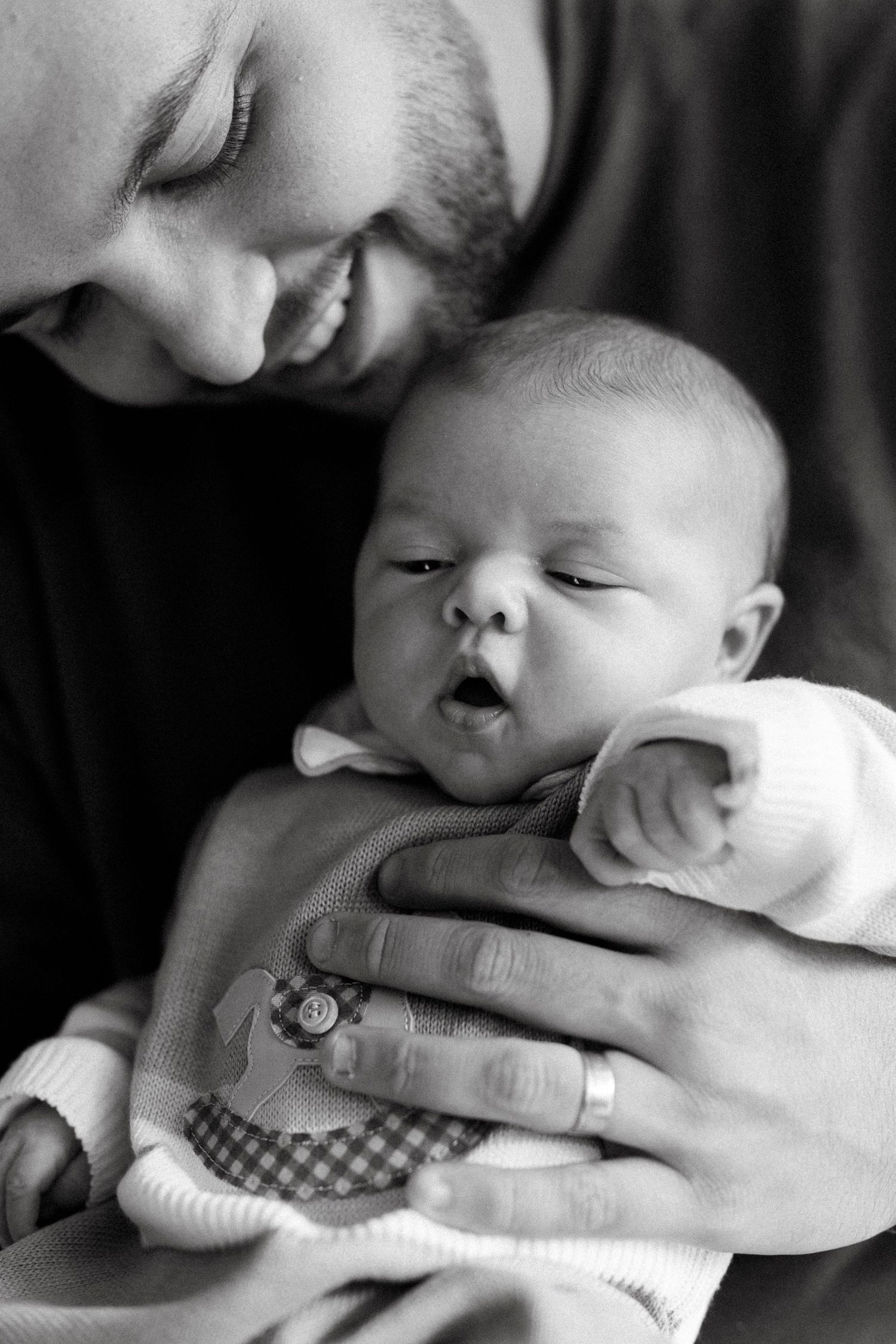 Black and white photo of a smiling father cradling his newborn baby, wearing a wedding ring.