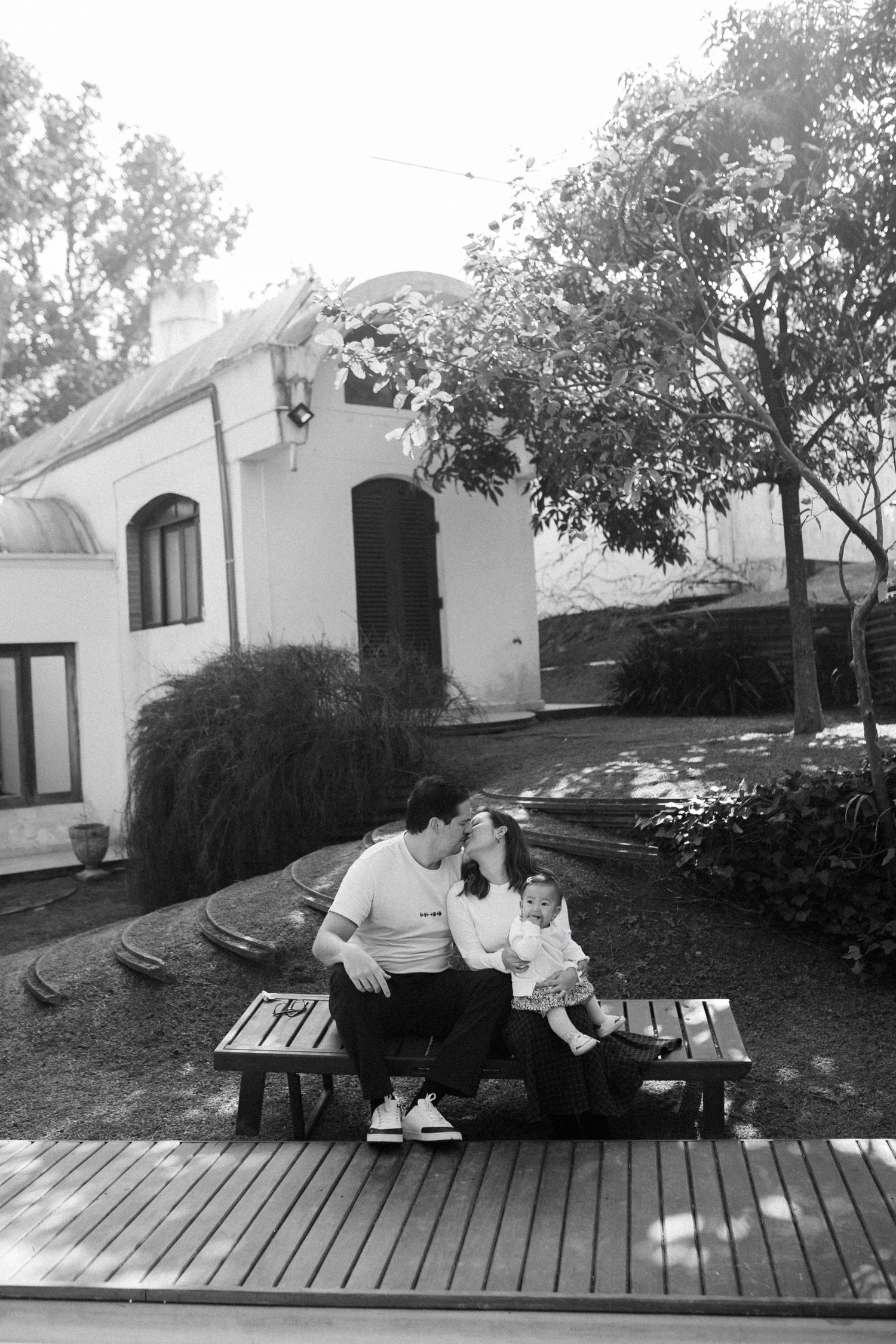 Black and white photo of a family sitting on a bench in a garden with a white house and trees in the background.