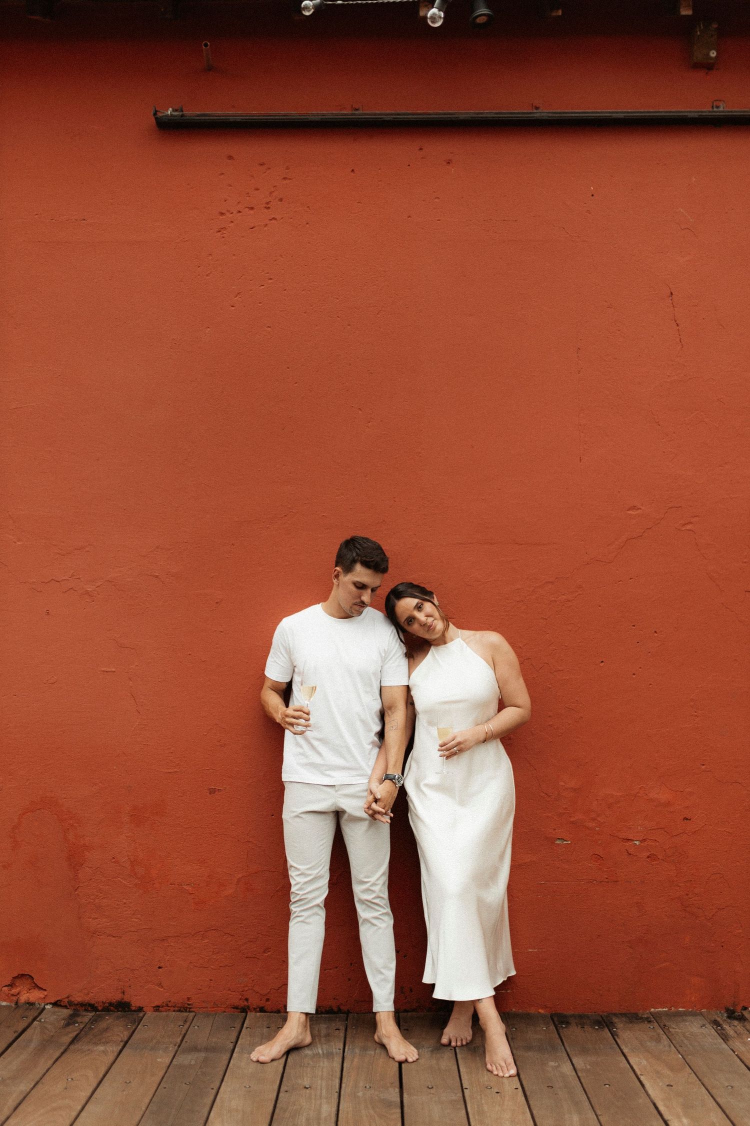 Couple in white outfits posing against a rustic red wall in an artistic photo sequence.