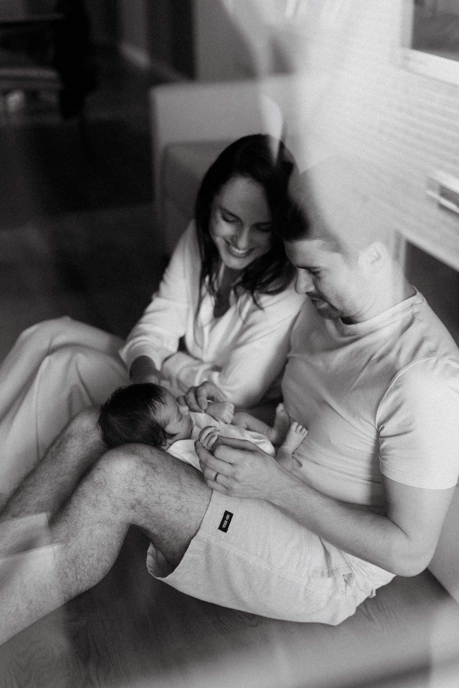 Black and white photo of smiling parents sitting together, lovingly cradling their newborn baby.
