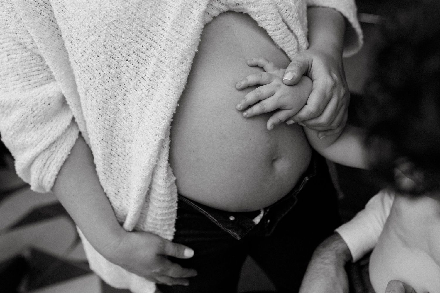 Black and white photo of a pregnant woman cradling her bare baby bump, with a partner's hand gently touching her belly.